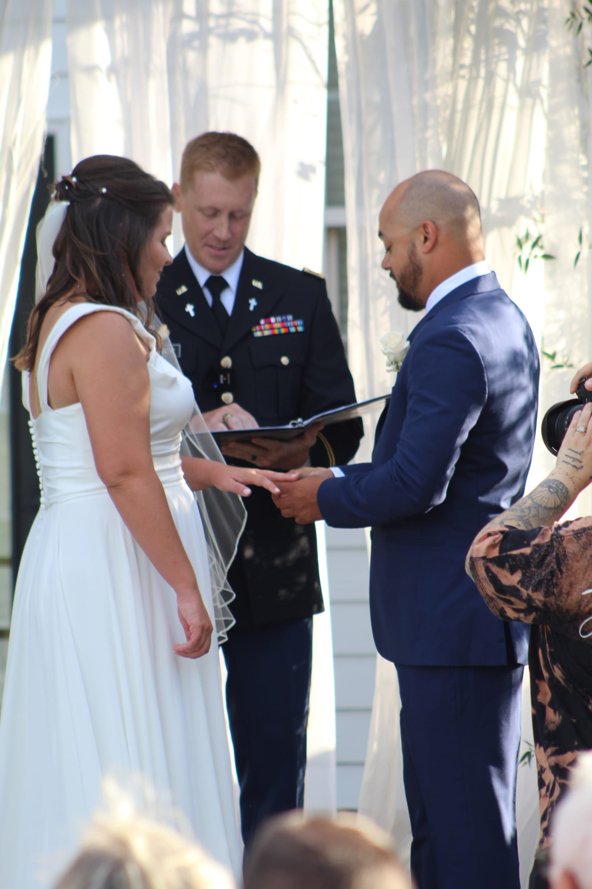 Bride in white gown and groom in navy suit exchange rings during an outdoor wedding ceremony officiated by a military officer at Scarborough House historic estate.