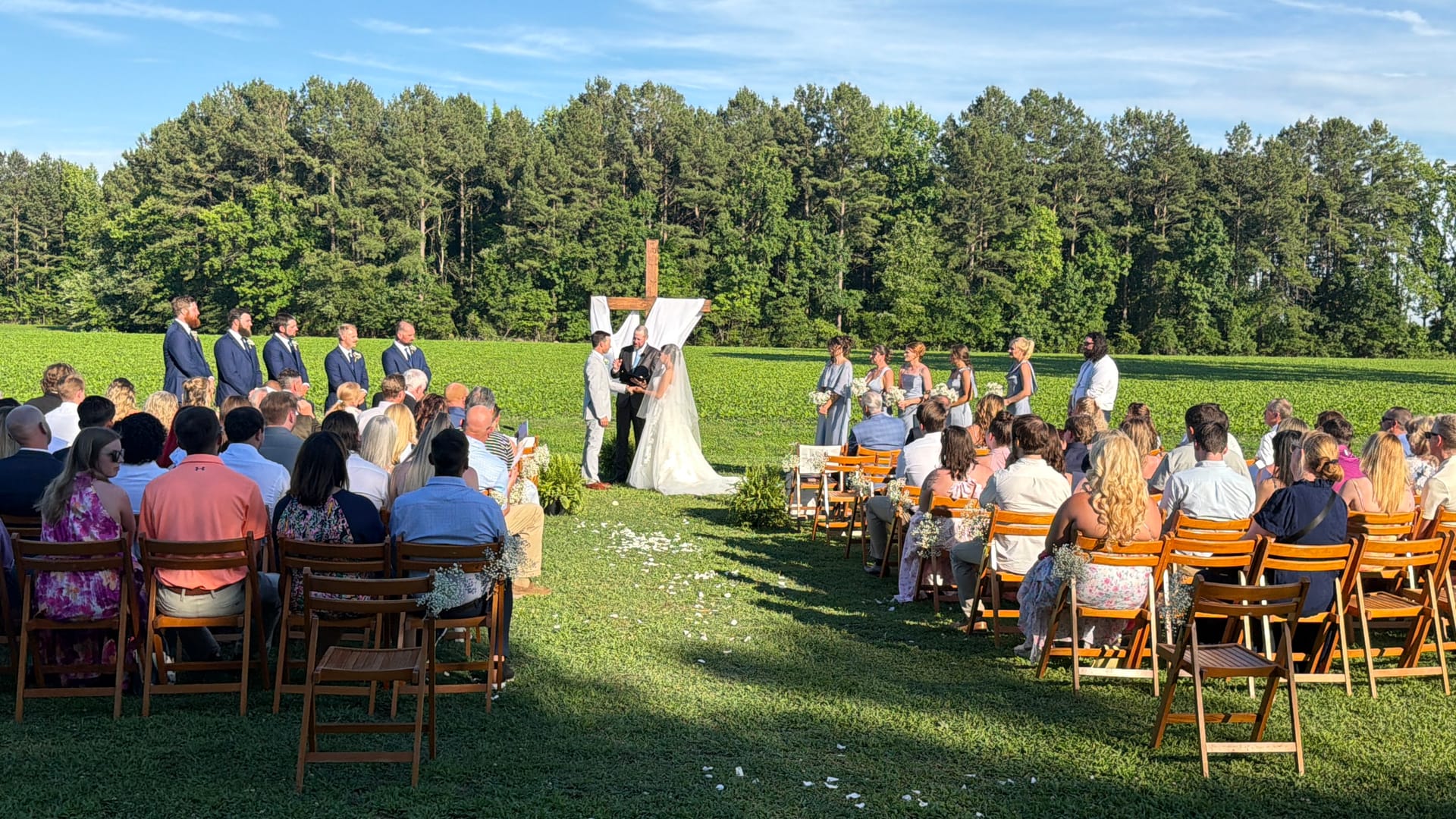 Outdoor wedding ceremony on the ceremony lawn at Scarborough House with guests seated in wooden chairs facing the couple at a rustic wooden arch backed by tall pine trees and green fields.
