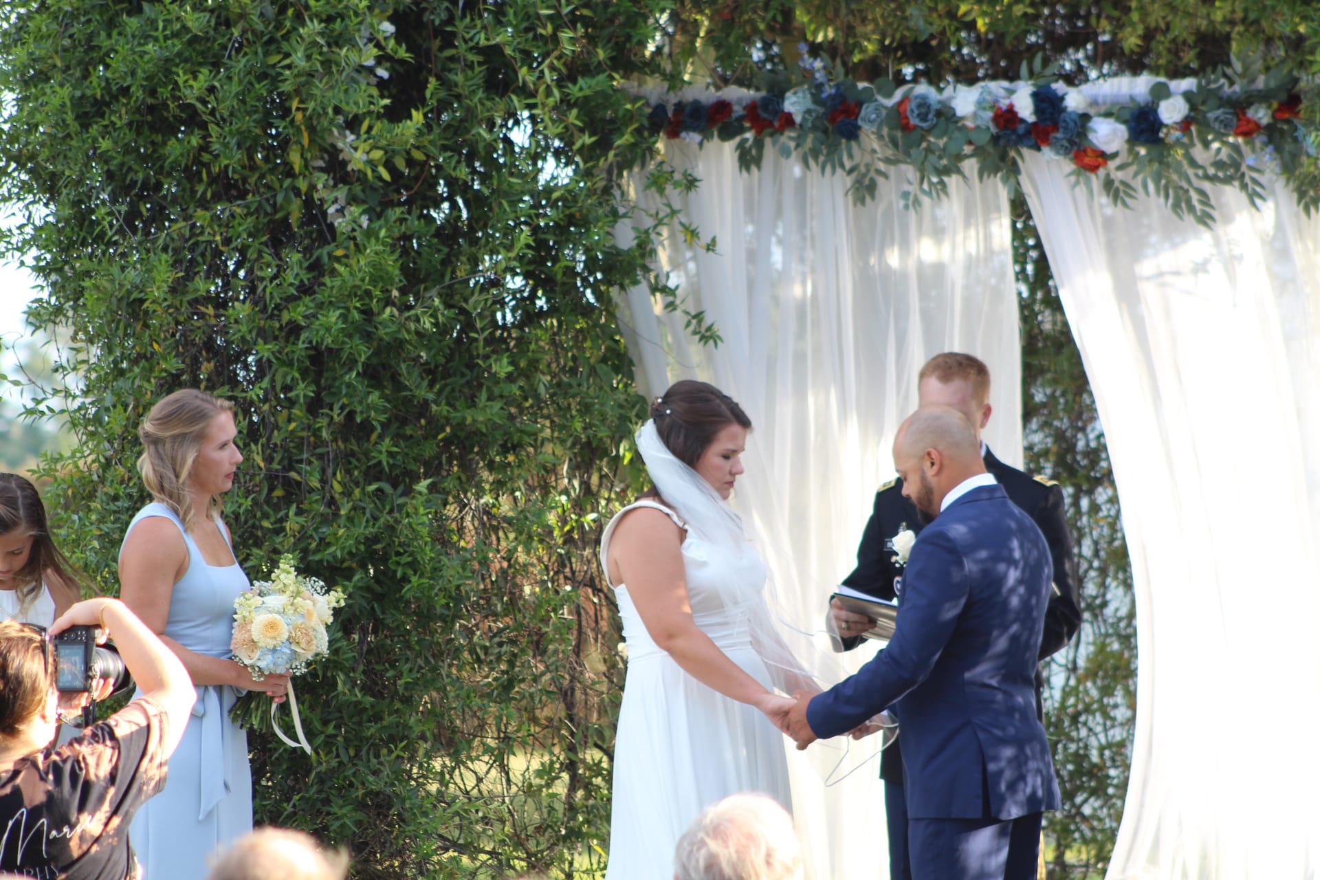 Bride and groom holding hands beneath a white draped floral arch adorned with navy and red roses during an outdoor wedding ceremony at Scarborough House.