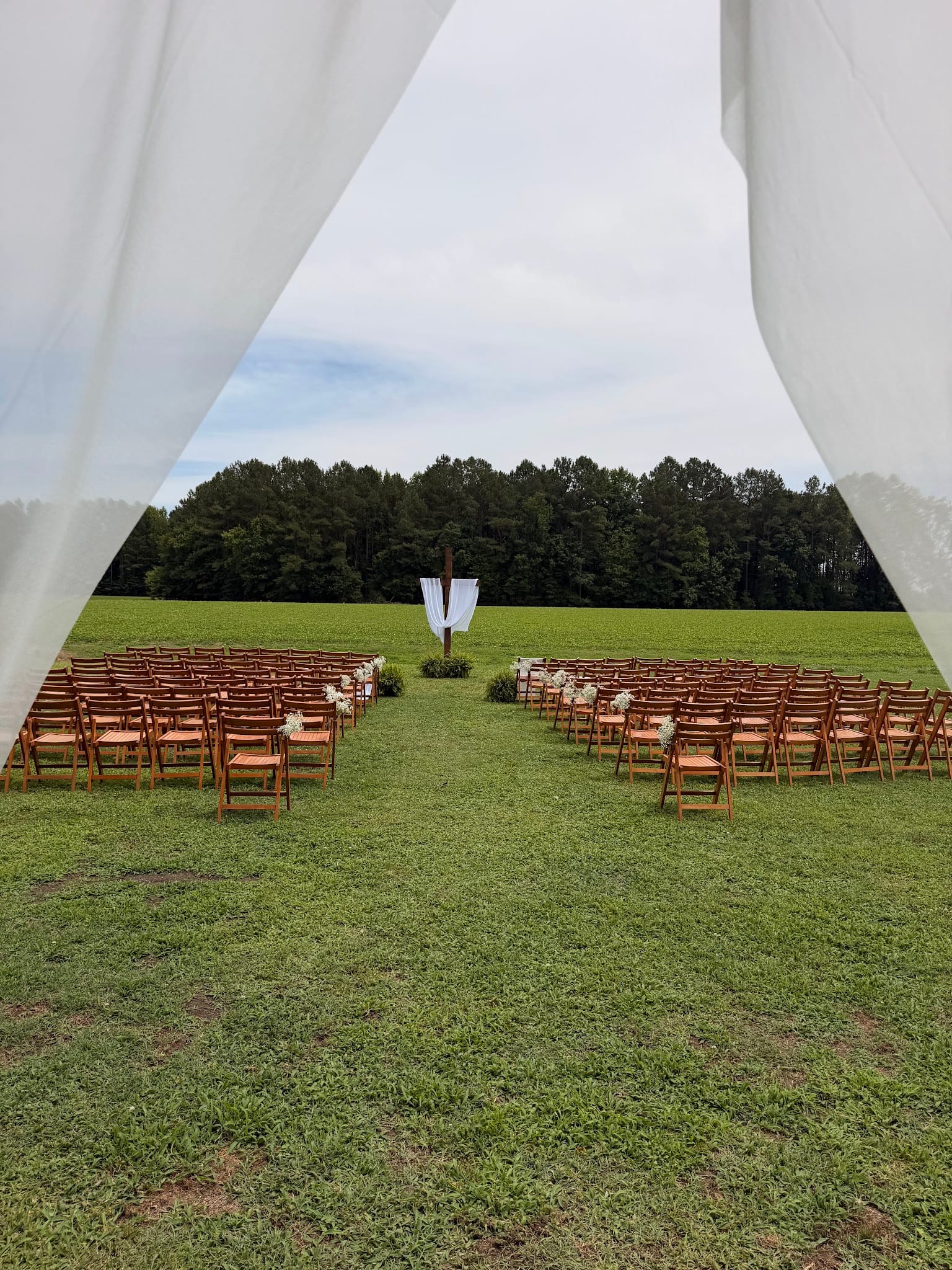 Outdoor wedding ceremony setup at Scarborough House viewed through white draping curtains, featuring rows of wooden folding chairs separated by a center aisle leading to a draped wooden arch on a lush green lawn with a wooded backdrop.