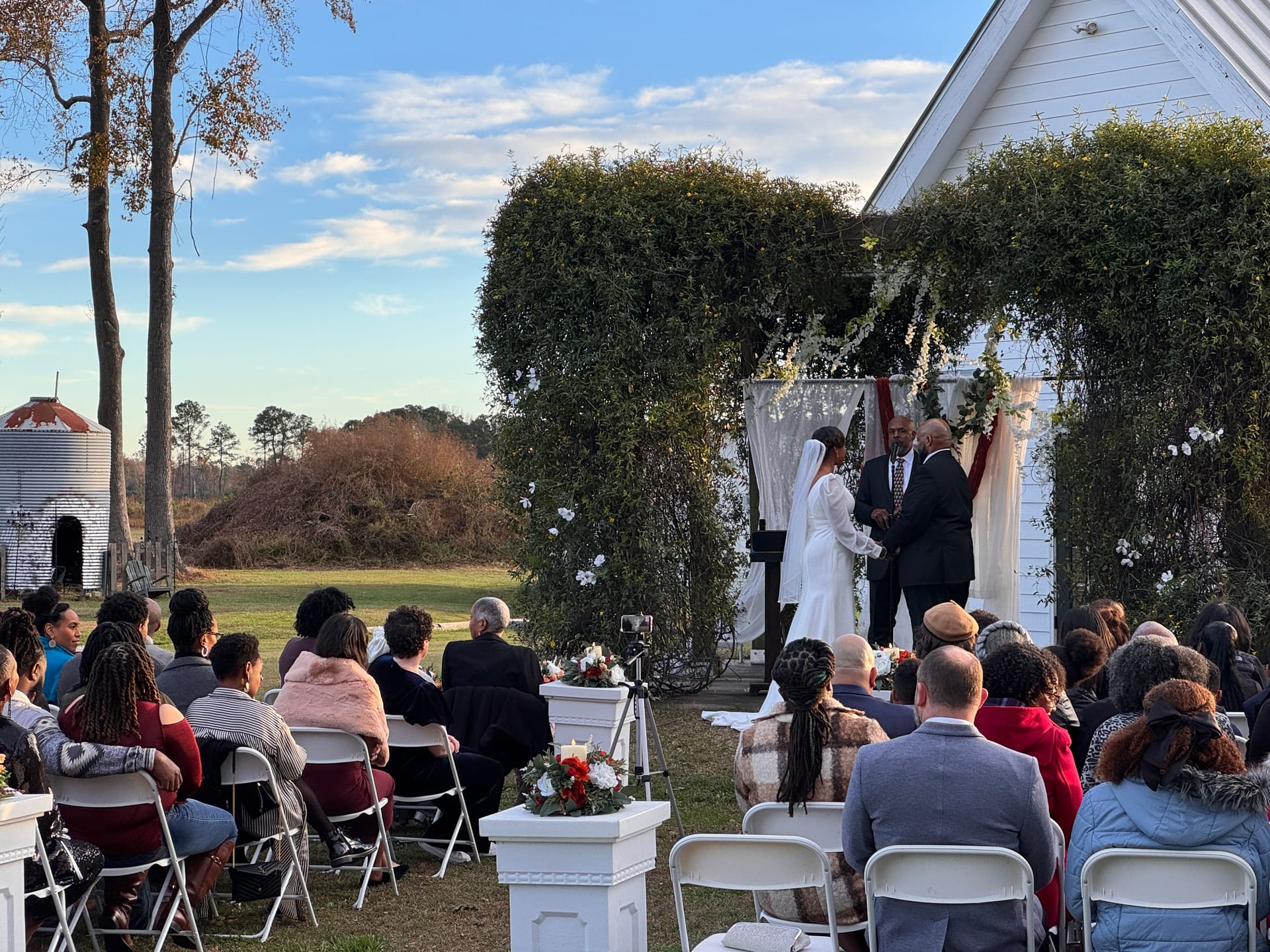 Bride and groom exchange vows beneath a lush greenery arch on the ceremony lawn at Scarborough House historic estate, surrounded by seated wedding guests on a sunny autumn afternoon.