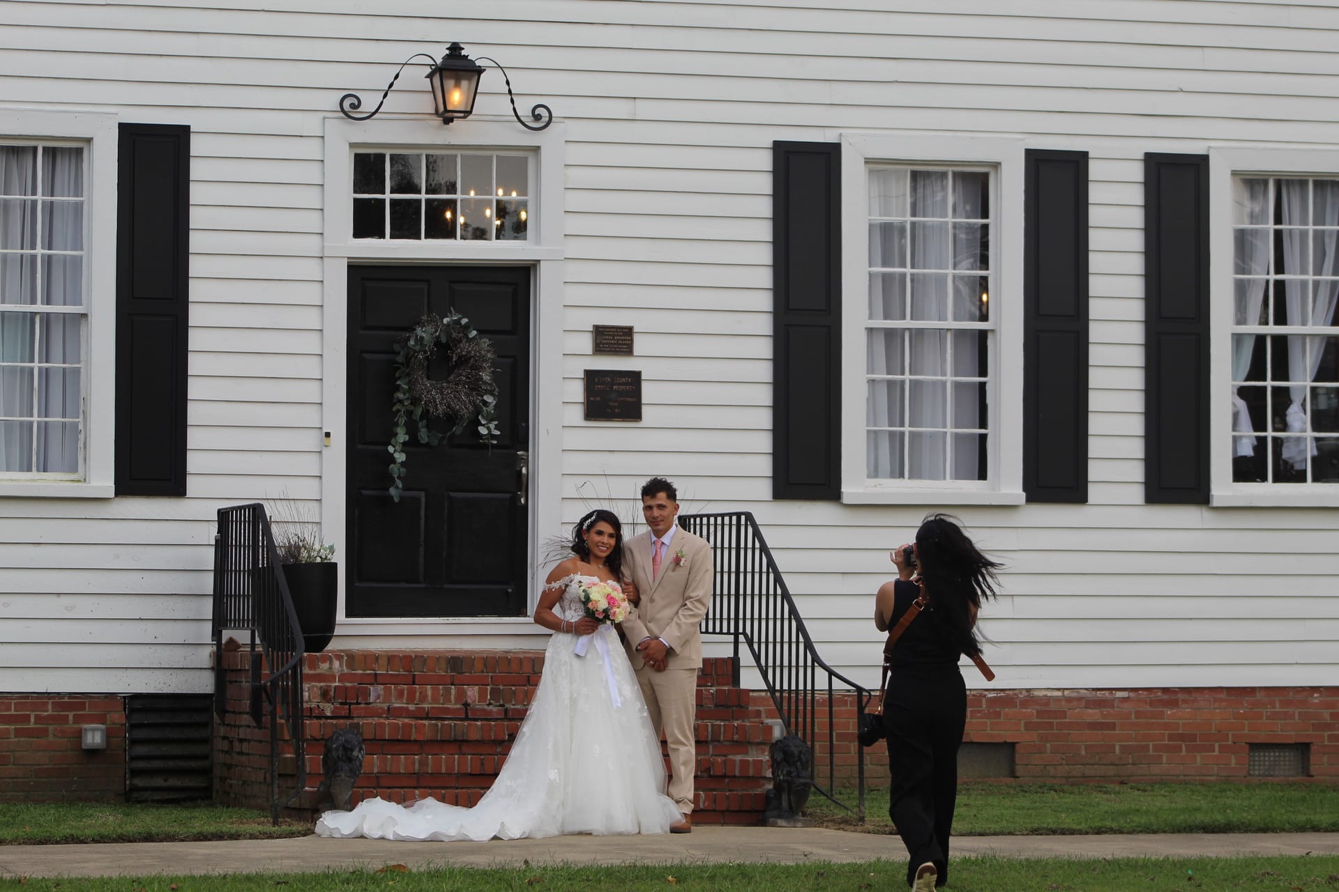 Bride in a white ball gown and groom in a tan suit pose together on the brick front steps of the historic 1821 Scarborough House manor while a photographer captures their wedding portrait.