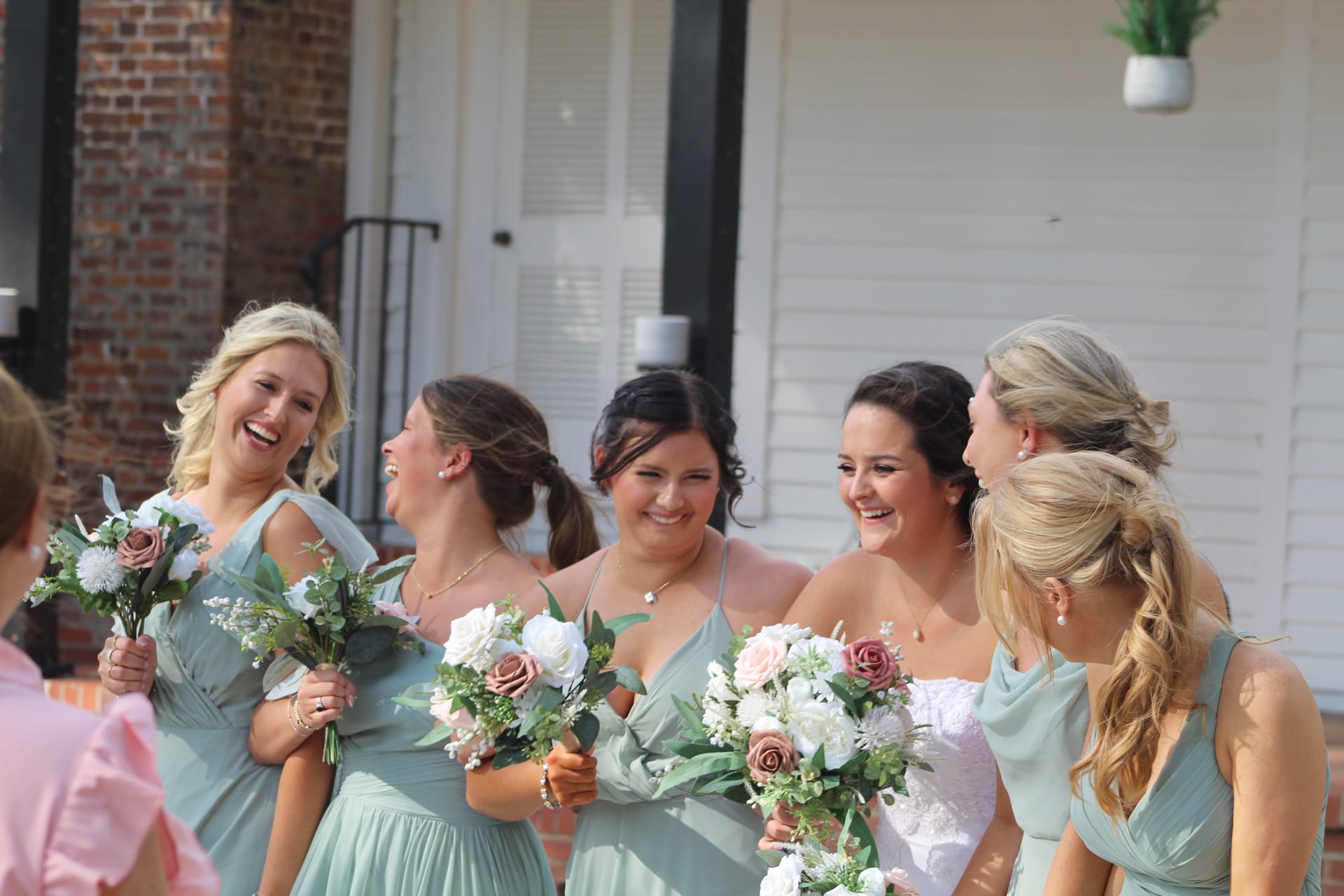 Bridesmaids in sage green gowns laugh joyfully with the bride in white lace while holding blush and ivory bouquets outside the historic white manor at Scarborough House.