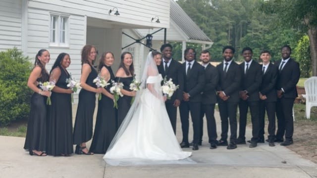 Bride in a white gown and veil stands centered between bridesmaids in black dresses and groomsmen in black suits outside the historic Scarborough House estate.