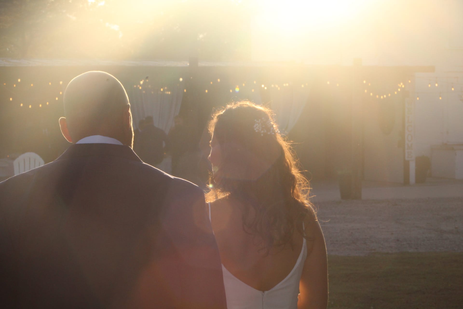 Bride and groom silhouetted against glowing golden sunset with string lights and the historic Scarborough House estate in the background.