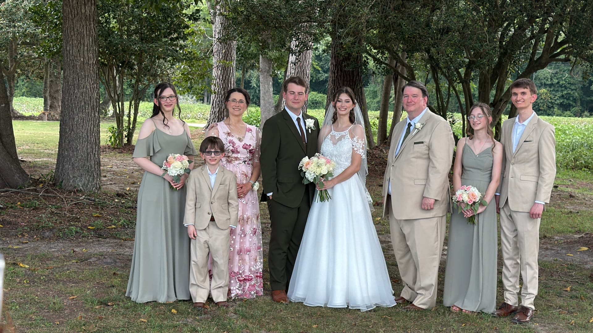 Bride and groom pose with their wedding party and family members in sage and tan formal attire beneath tall oak trees at Scarborough House estate.