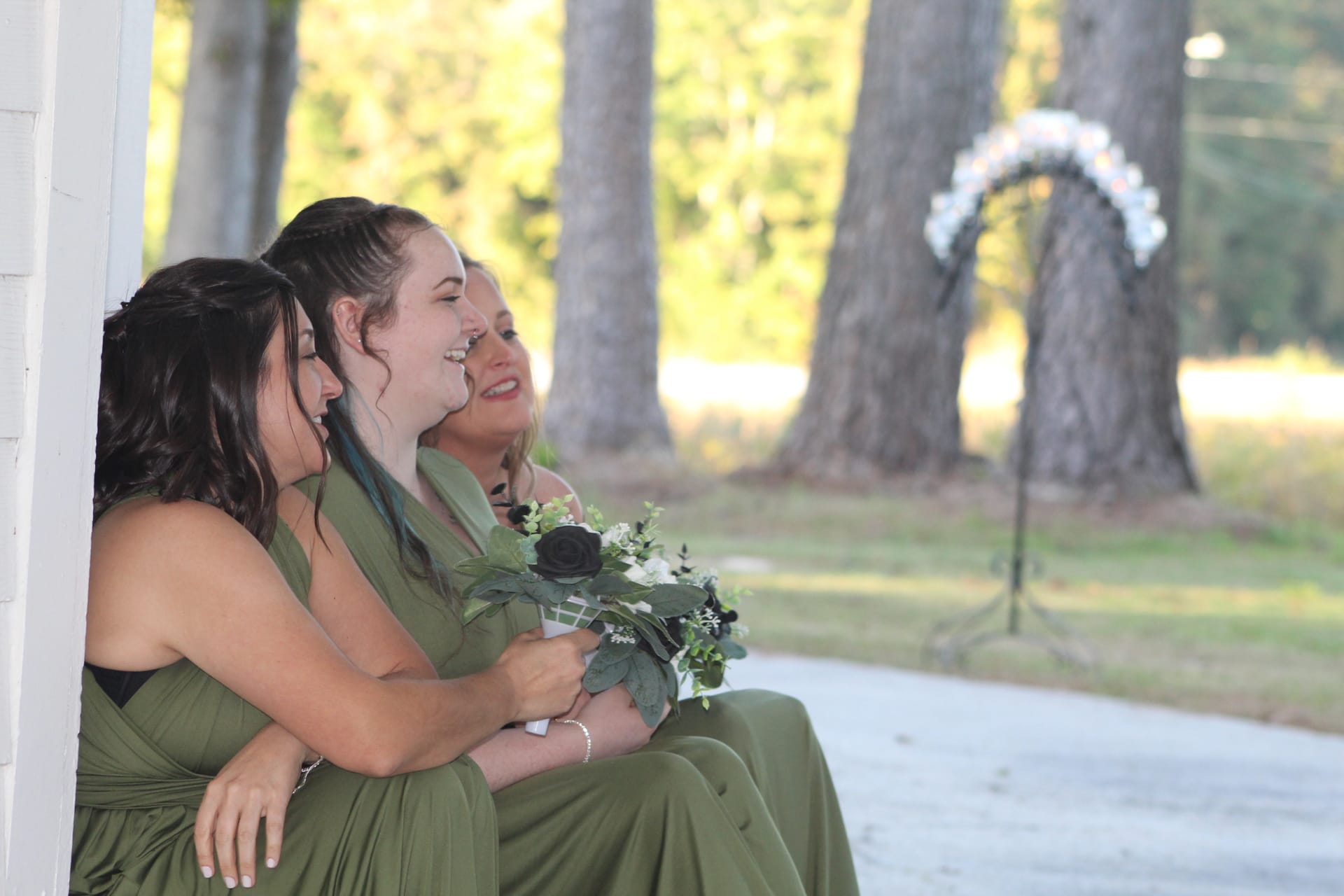 Three bridesmaids in sage green dresses laughing together on the porch at Scarborough House with a floral arch and towering oak trees in the background.
