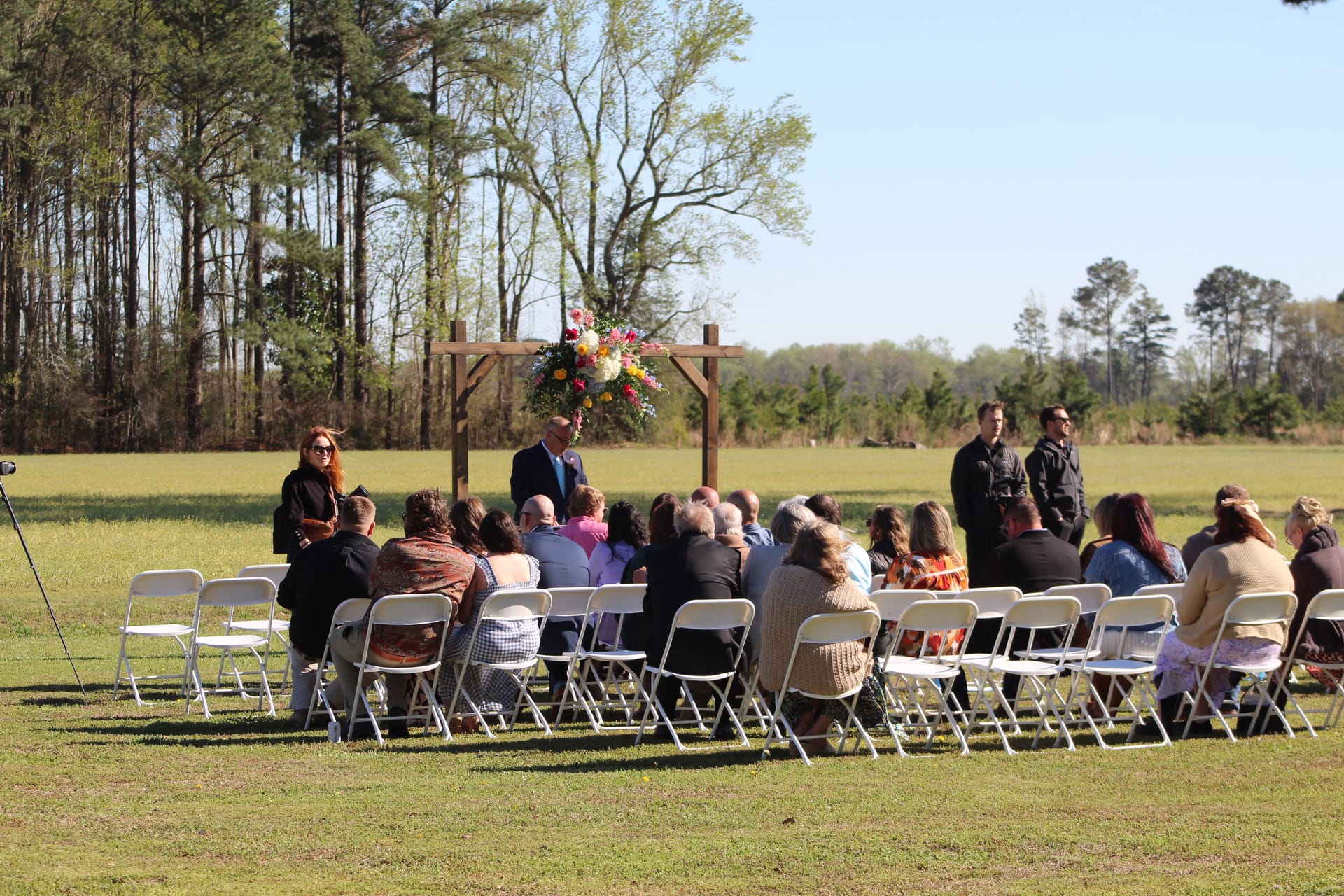 Guests seated in white folding chairs during an outdoor wedding ceremony beneath a wooden arch adorned with colorful florals on the open ceremony lawn at Scarborough House historic estate.