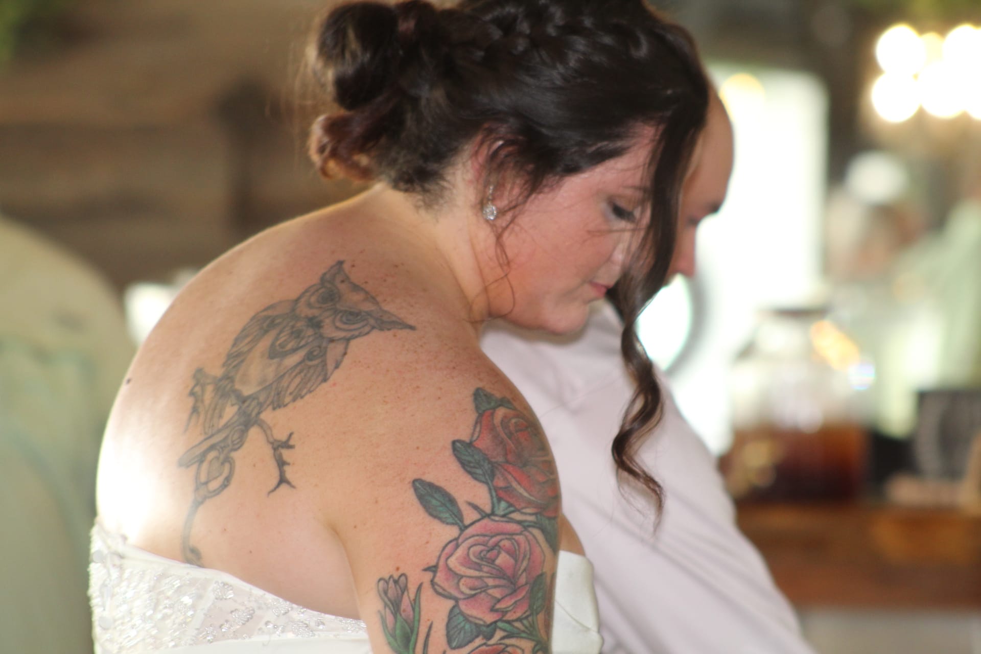 Bride in white strapless wedding gown with an intricate owl and rose tattoo sleeve displayed on her shoulder as she looks down during wedding preparations.