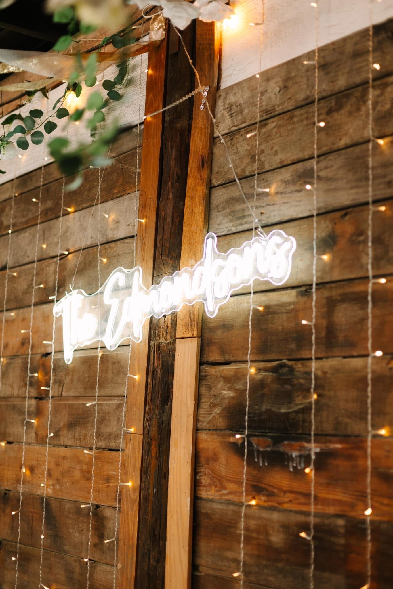 A glowing white neon sign reading The Shandons mounted on a rustic reclaimed wood wall adorned with cascading fairy lights and eucalyptus greenery at Scarborough House wedding venue.