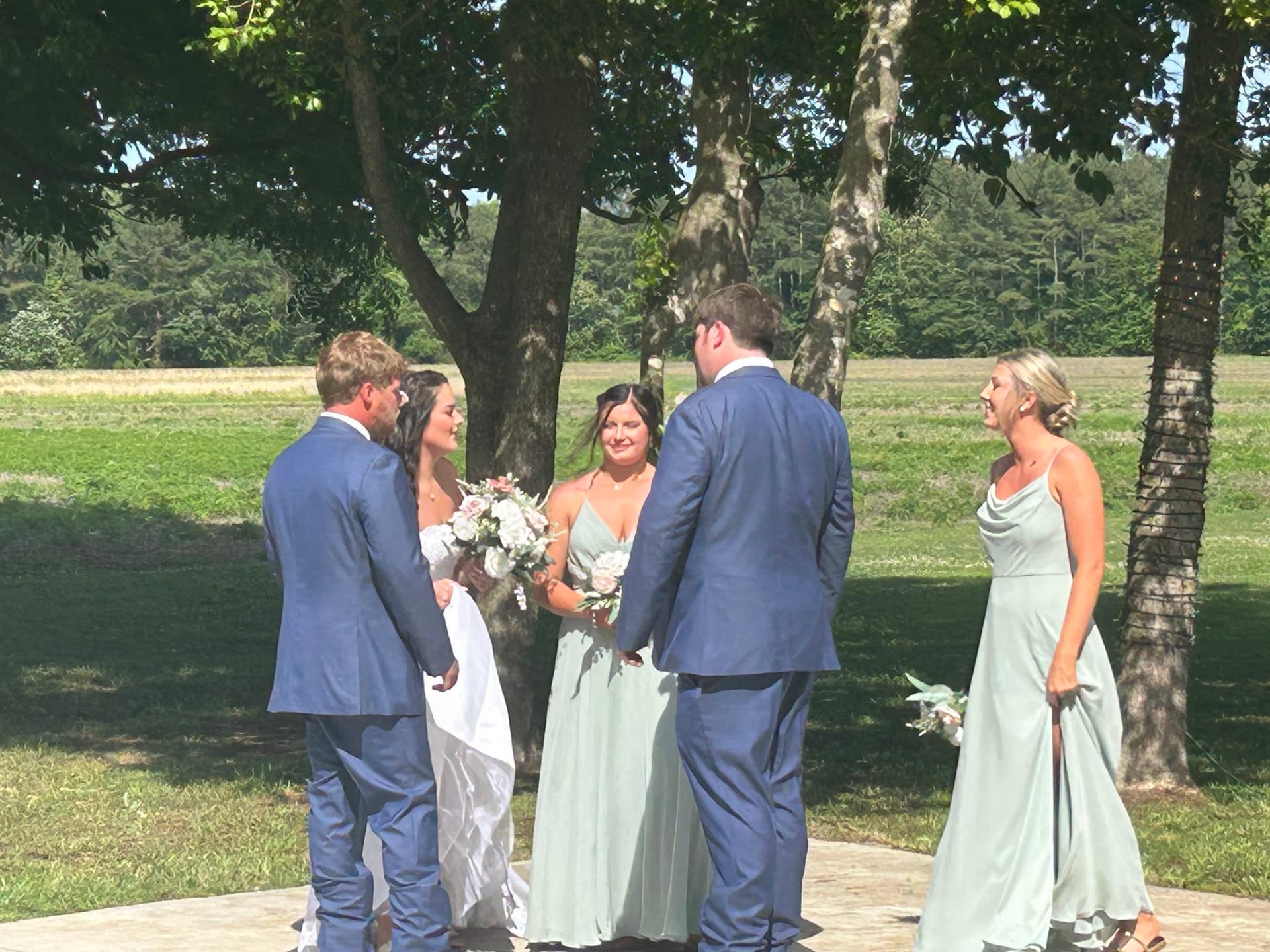 Bride and groom exchange vows outdoors beneath oak trees surrounded by bridesmaids in sage green gowns and groomsmen in navy suits on the ceremony lawn at Scarborough House.