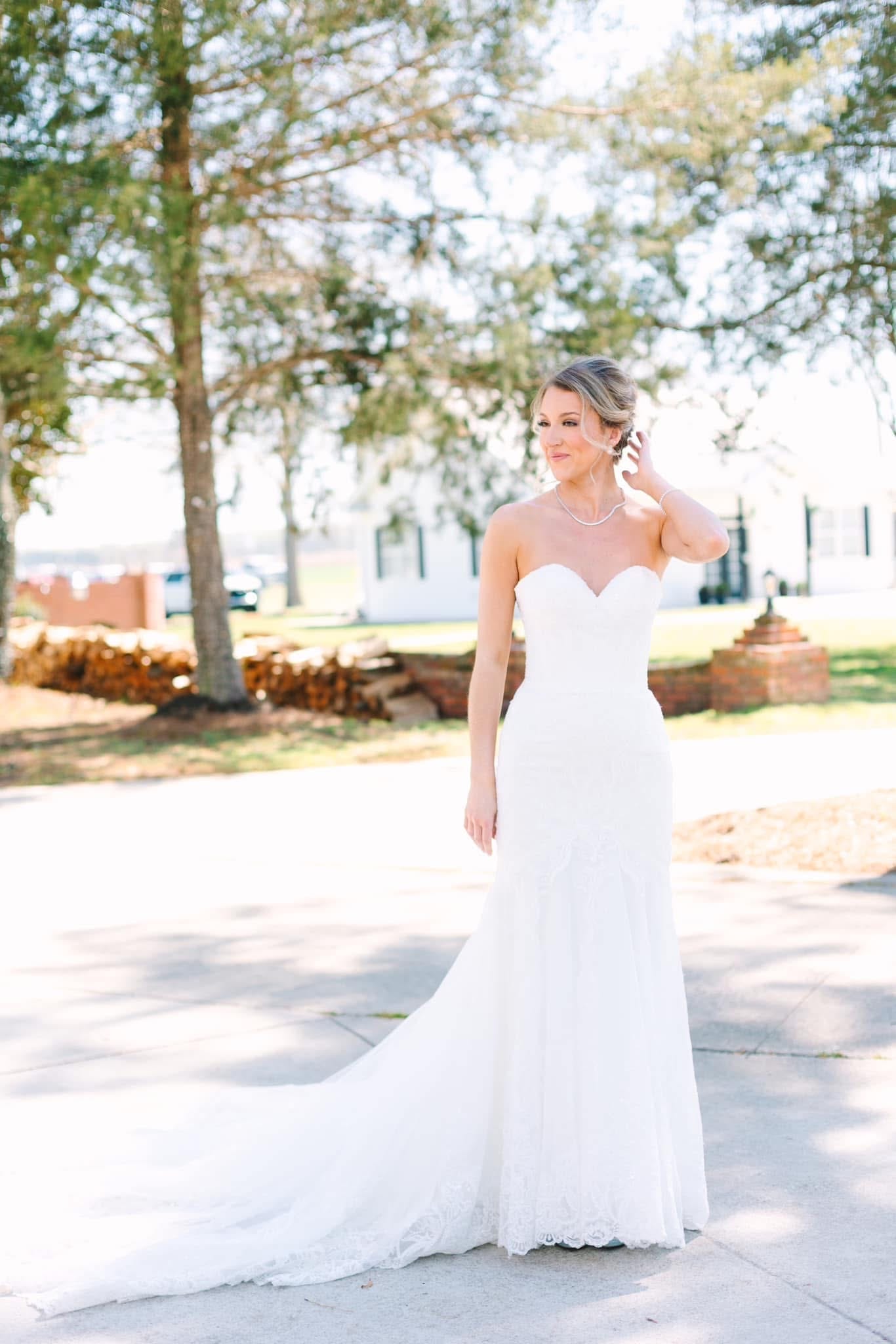 Bride in a strapless sweetheart lace wedding gown with cathedral train poses gracefully on the sunlit grounds of Scarborough House historic estate surrounded by mature trees.