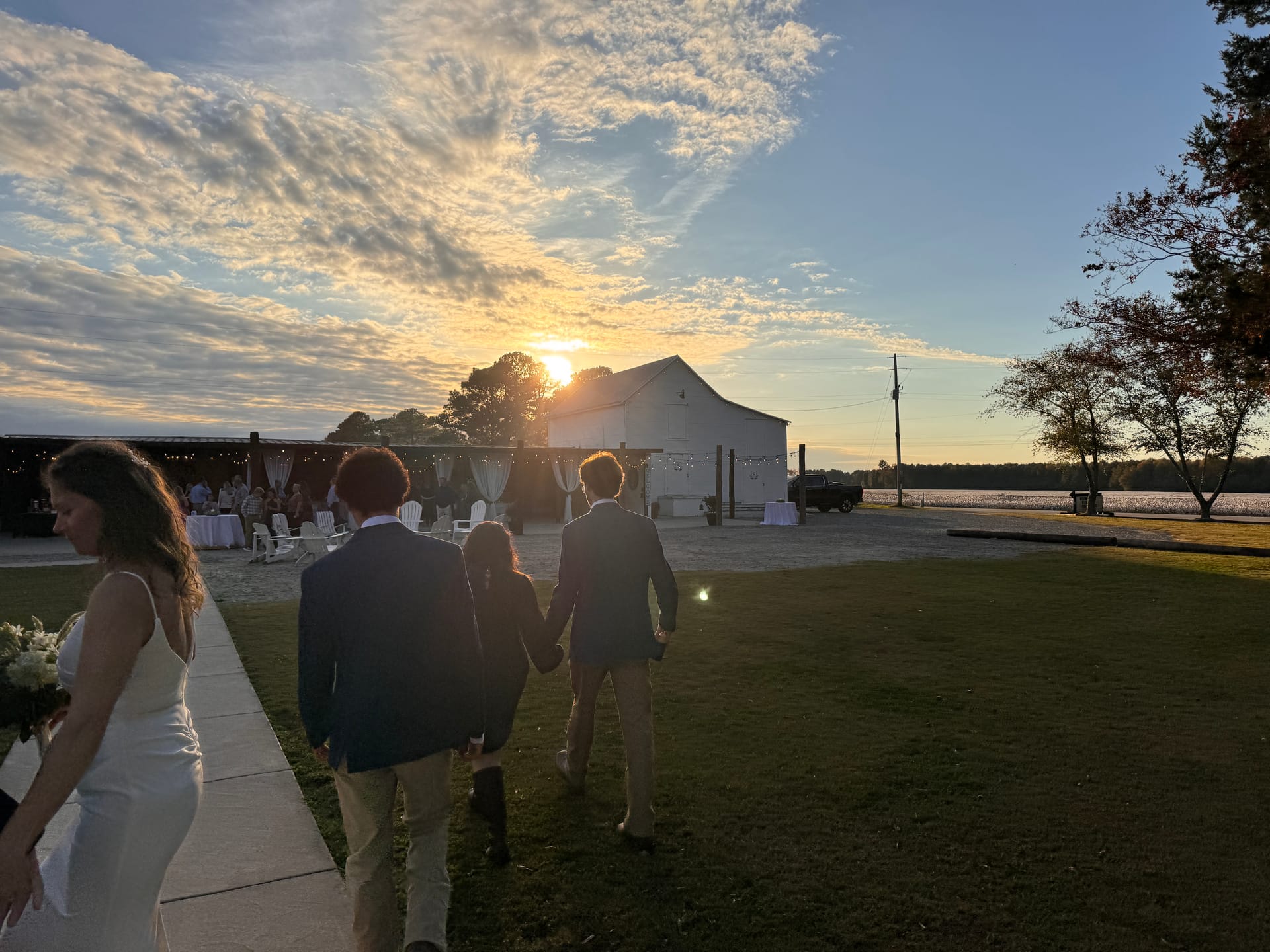 Wedding party silhouetted at sunset walking toward a white barn venue beside a lake with a dramatic golden sky.