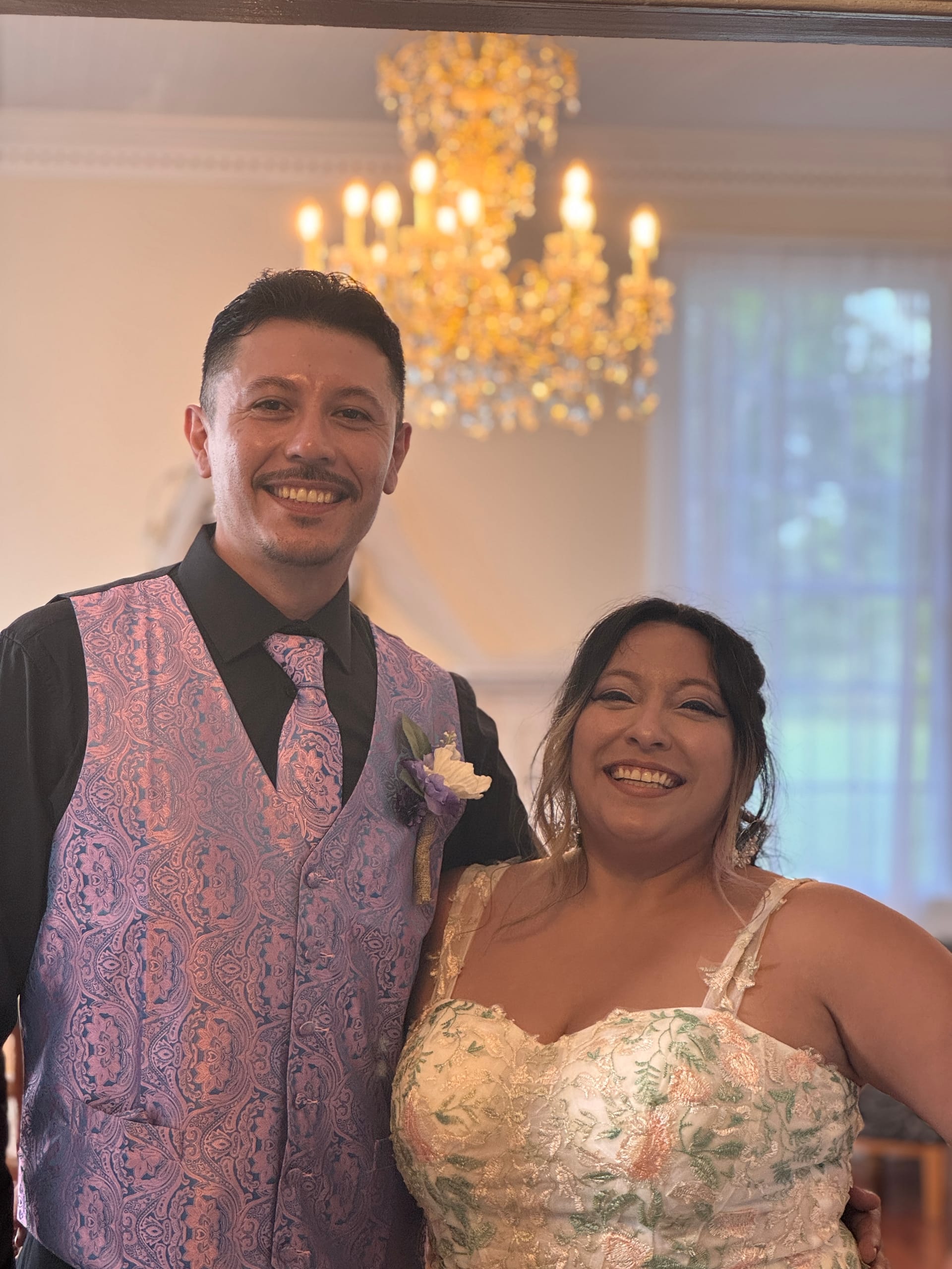 Smiling bride in a floral lace gown and groom in a paisley vest and tie pose together beneath a golden chandelier inside the historic grand hall at Scarborough House.