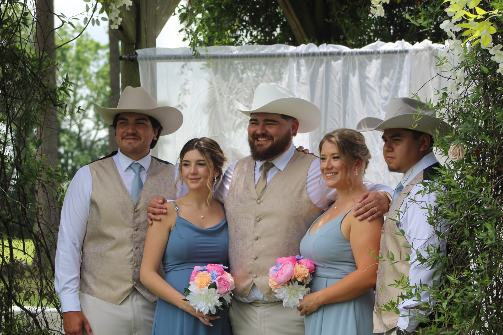 Groomsmen in tan vests and cowboy hats pose with bridesmaids in dusty blue dresses holding colorful bouquets beneath a floral archway at an outdoor wedding ceremony.