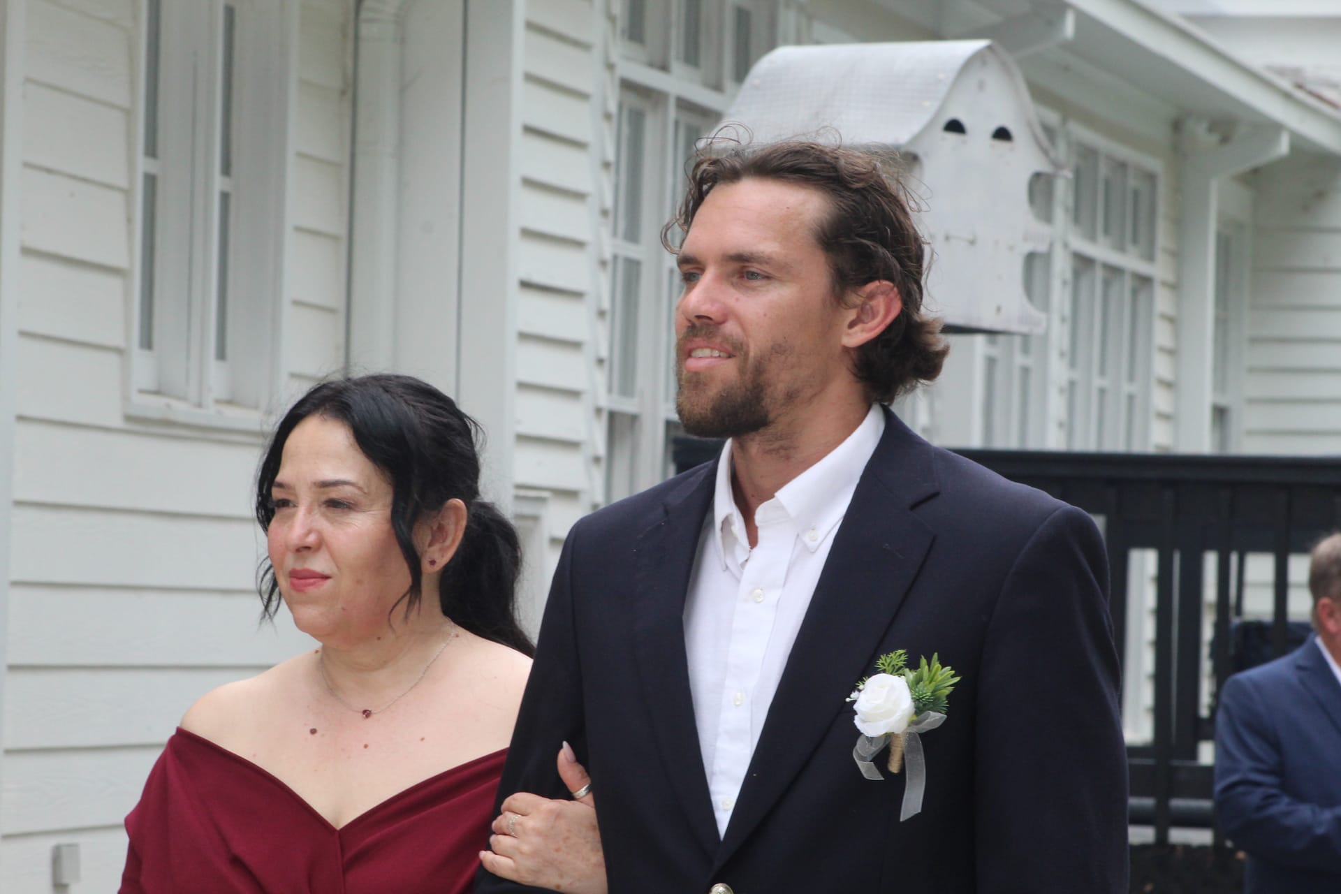 A groomsman in a navy suit with a white rose boutonniere escorts a bridesmaid in a burgundy off-shoulder gown along the historic white clapboard exterior of Scarborough House.