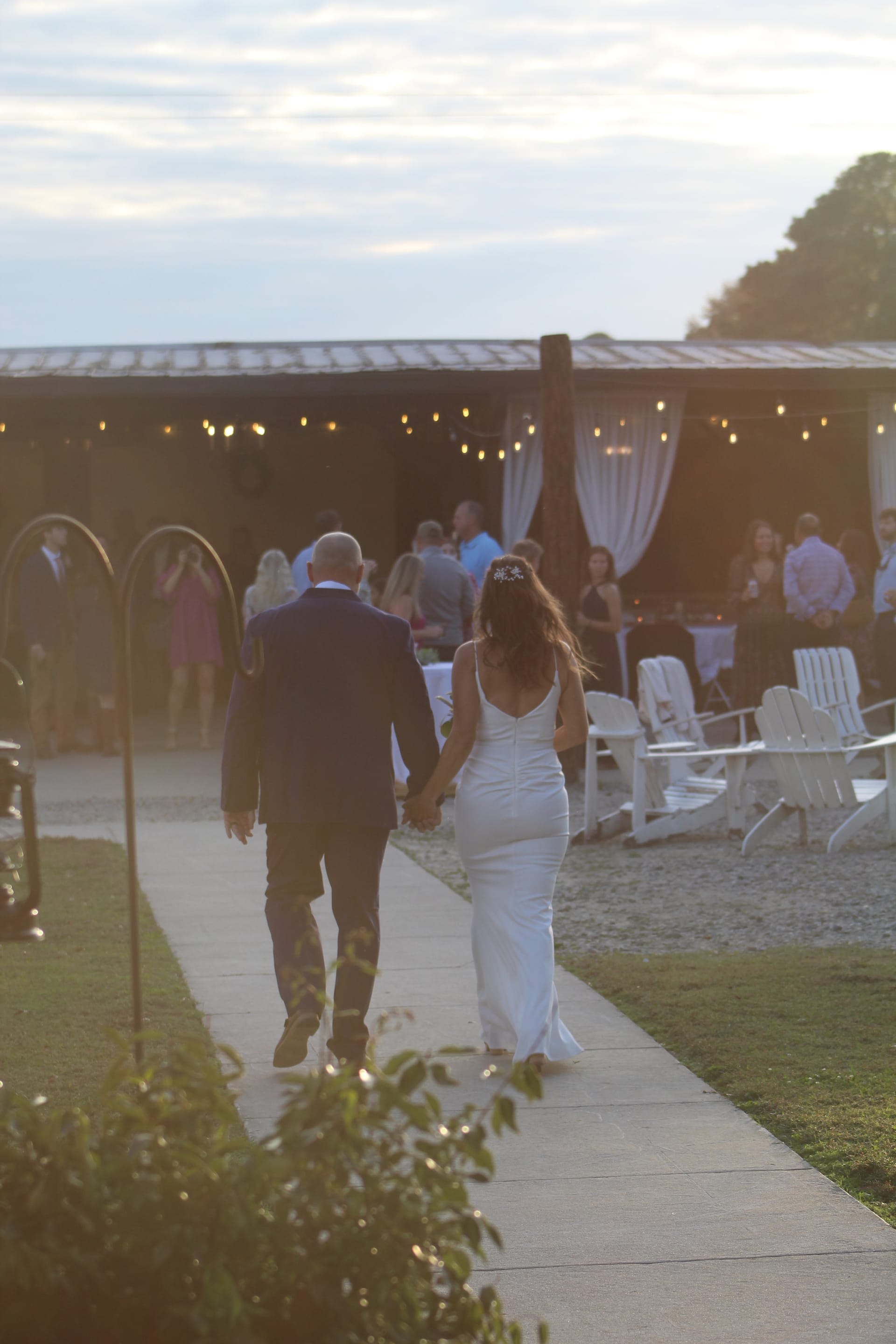 Bride in a fitted white gown and groom in a dark suit walk hand-in-hand along a pathway toward a string-light-adorned pavilion at golden hour during their Scarborough House wedding reception.