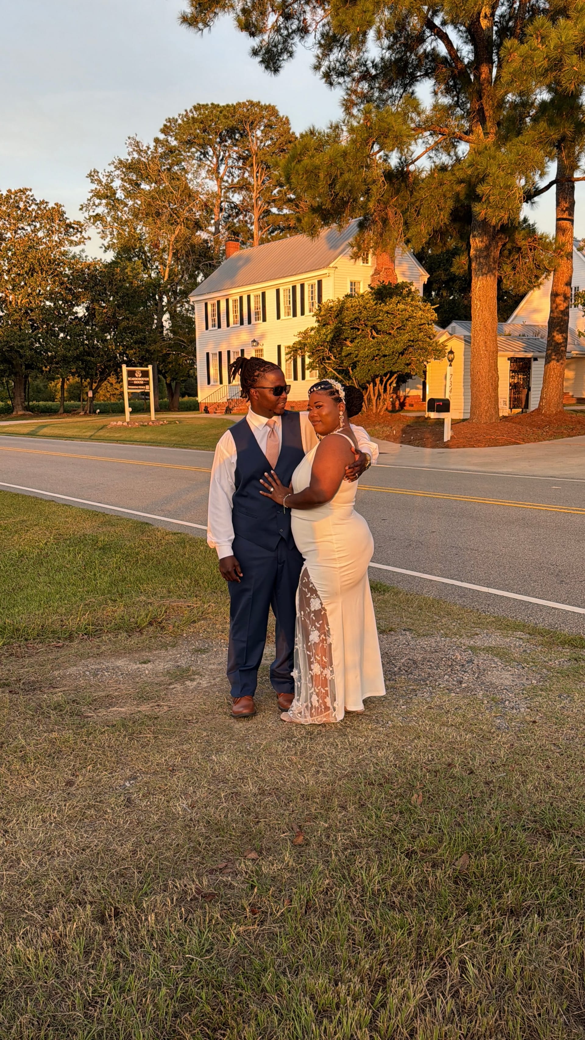 Bride in a white lace gown and groom in a navy vest and pink tie share a romantic embrace in golden evening light in front of the historic 1821 Scarborough House manor.