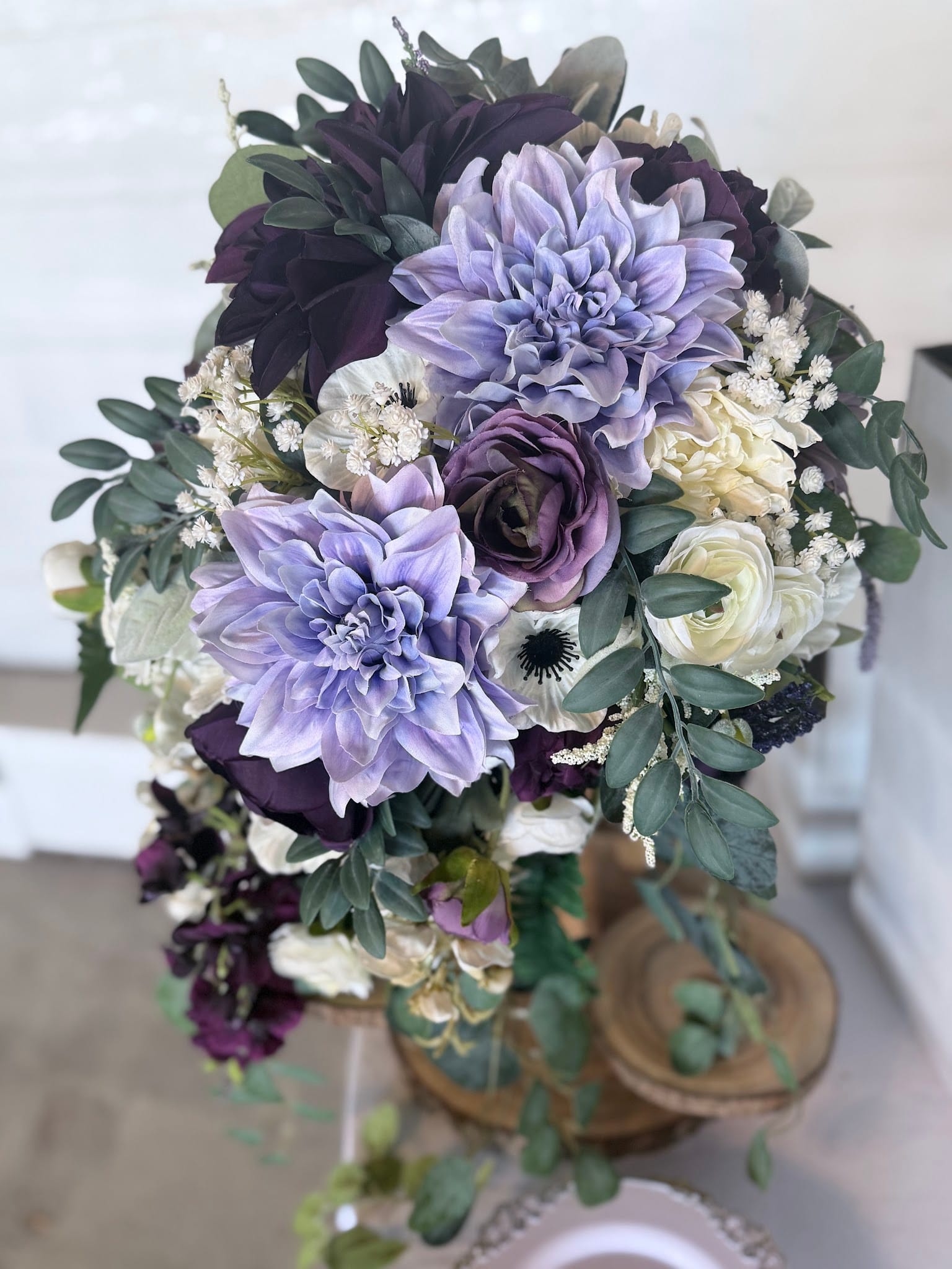 A lush cascading bridal bouquet featuring lavender dahlias, deep plum roses, white anemones, and trailing eucalyptus displayed on a rustic wooden stand at Scarborough House.