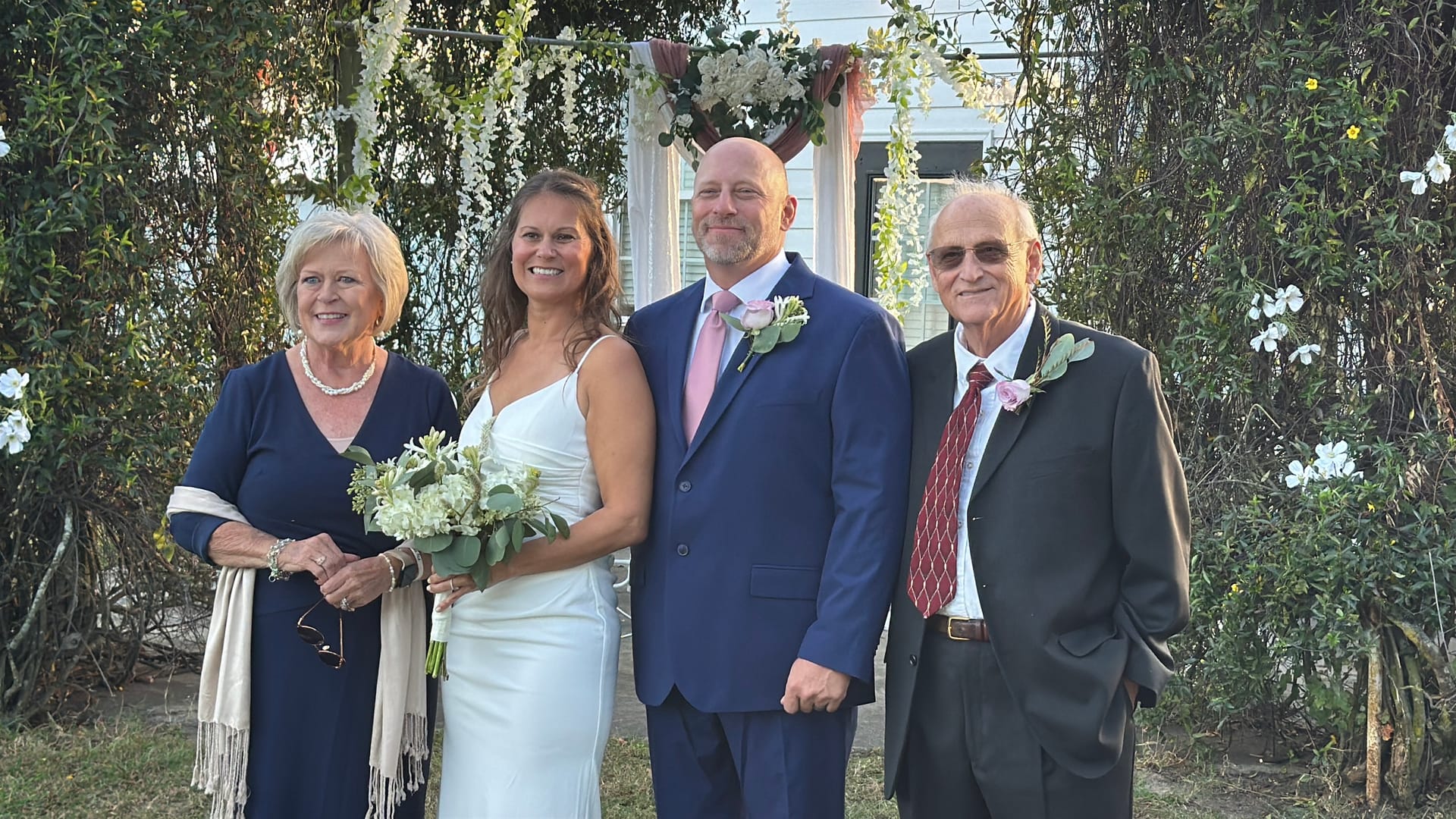 Bride in white gown holding a white floral bouquet stands with her groom in a navy suit and two family members beneath a floral arch surrounded by lush greenery at Scarborough House.
