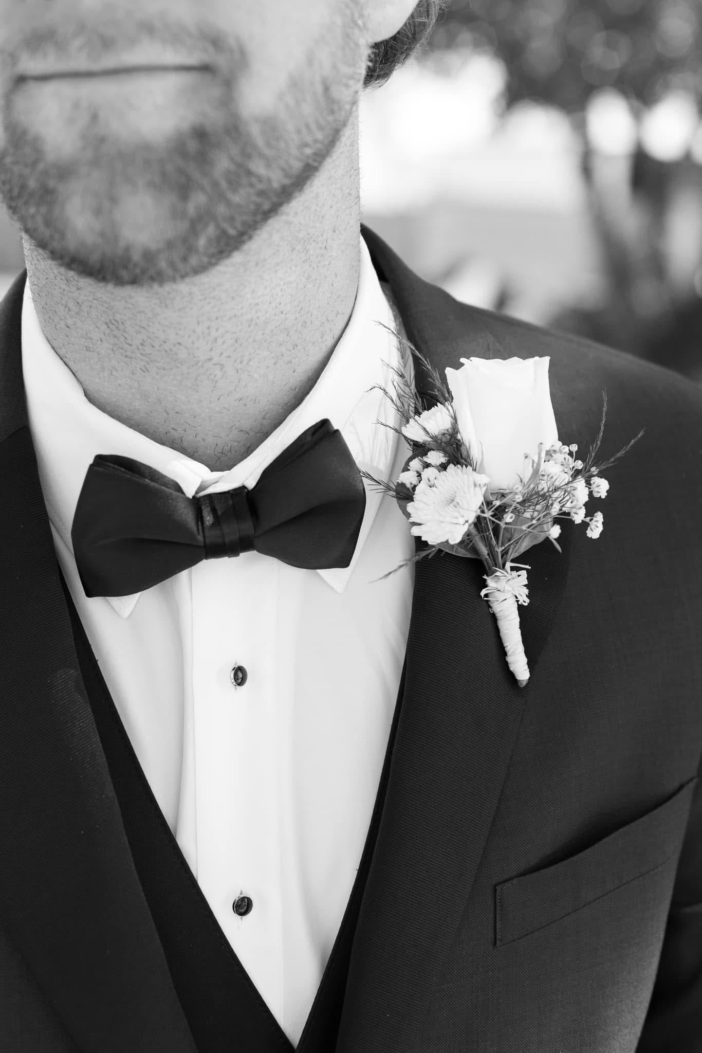 Black and white close-up of a groom in a classic tuxedo with a black bow tie and a floral boutonnière featuring a rose and baby's breath at a wedding.