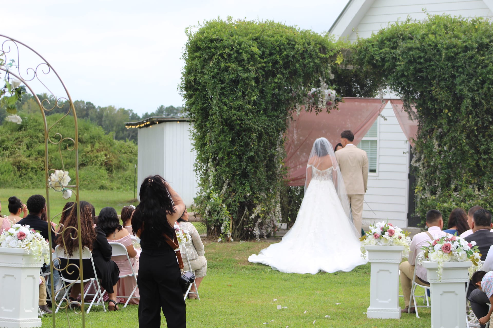 Bride in a white ball gown with cathedral veil walks toward groom at a lush ivy-covered ceremony arch adorned with mauve drapery and florals on Scarborough House ceremony lawn, flanked by white pedestal arrangements of pink and white flowers as guests look on.
