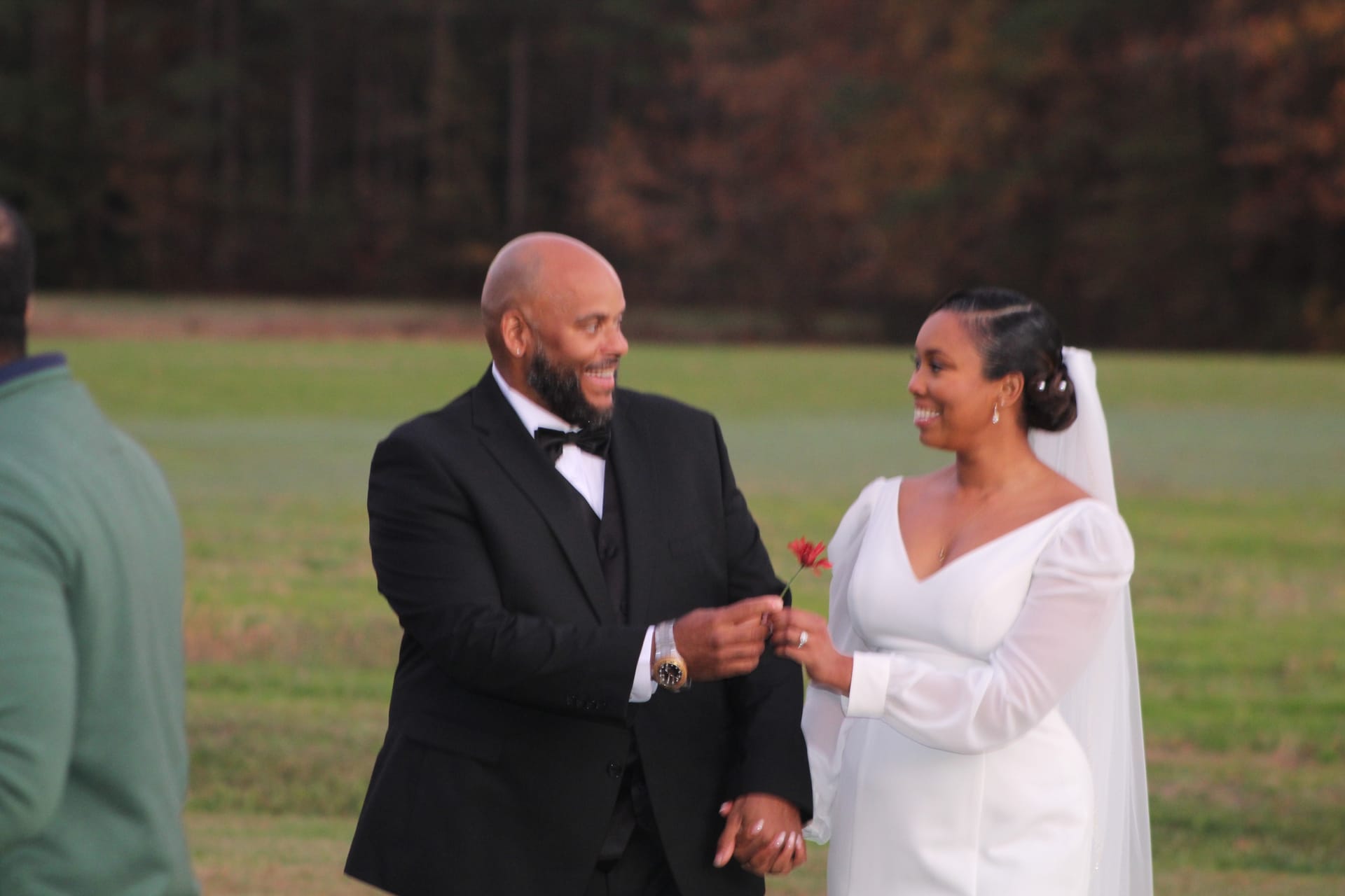 A smiling groom in a black tuxedo presents a red flower to his radiant bride in a long-sleeve white gown and veil during an outdoor autumn ceremony on the estate fields.