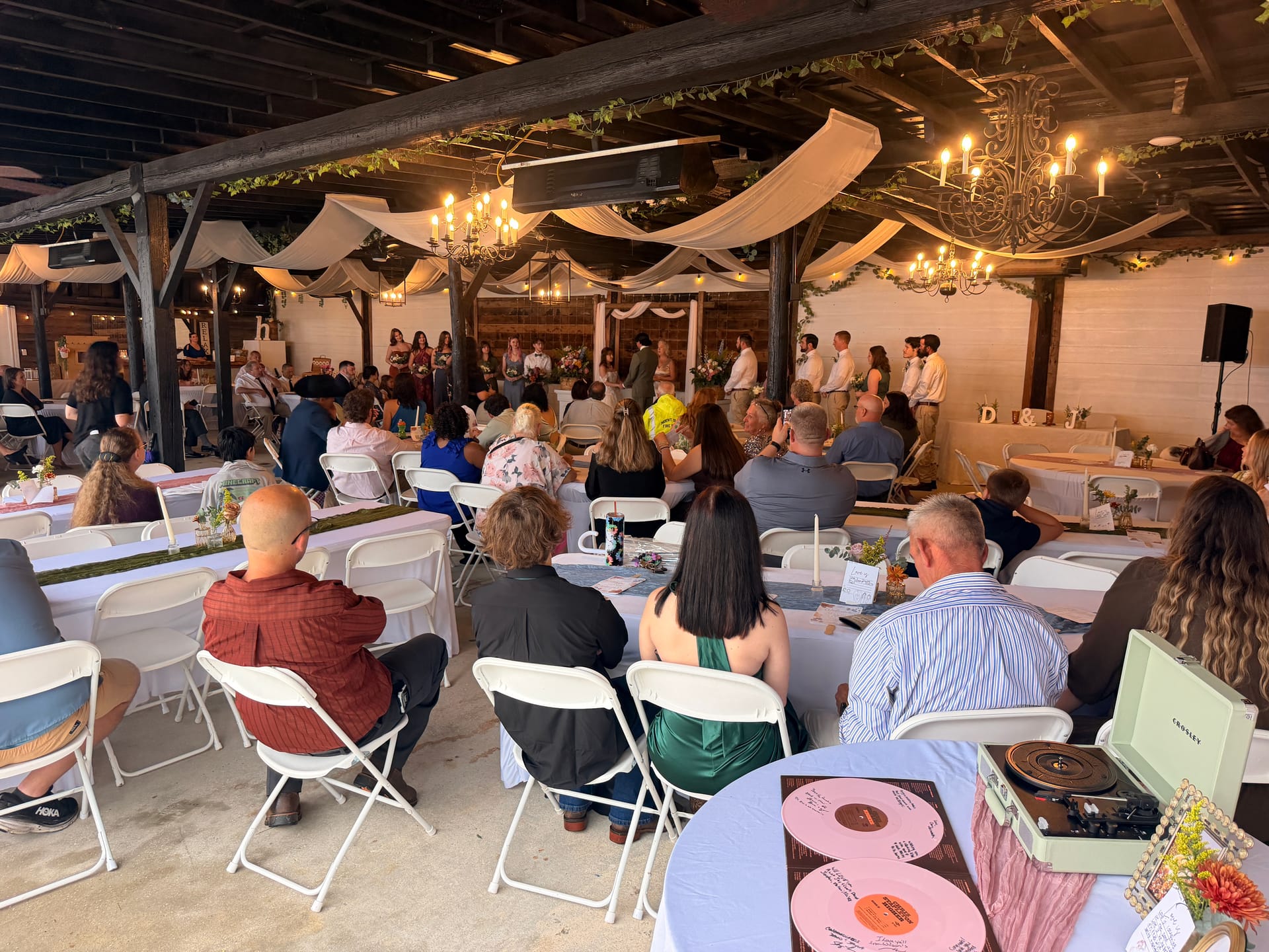 Wedding guests seated at round tables watch a ceremony in a warmly lit rustic pavilion adorned with draped fabric, chandeliers, and greenery while the bridal party stands at a floral altar in the background.
