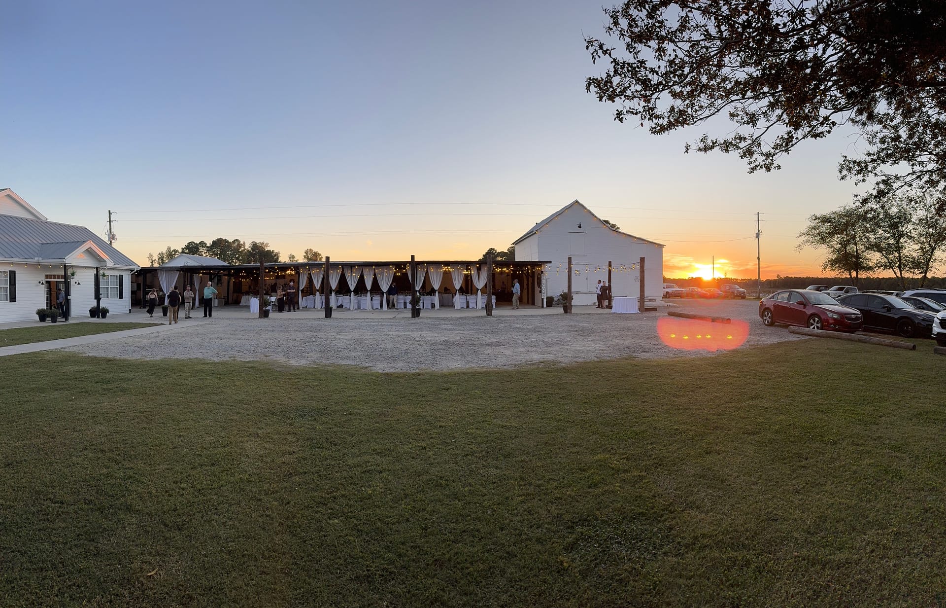 Panoramic sunset view of an elegant outdoor wedding reception at Scarborough House featuring a draped open-air pavilion with string lights and white barn against a golden evening sky.