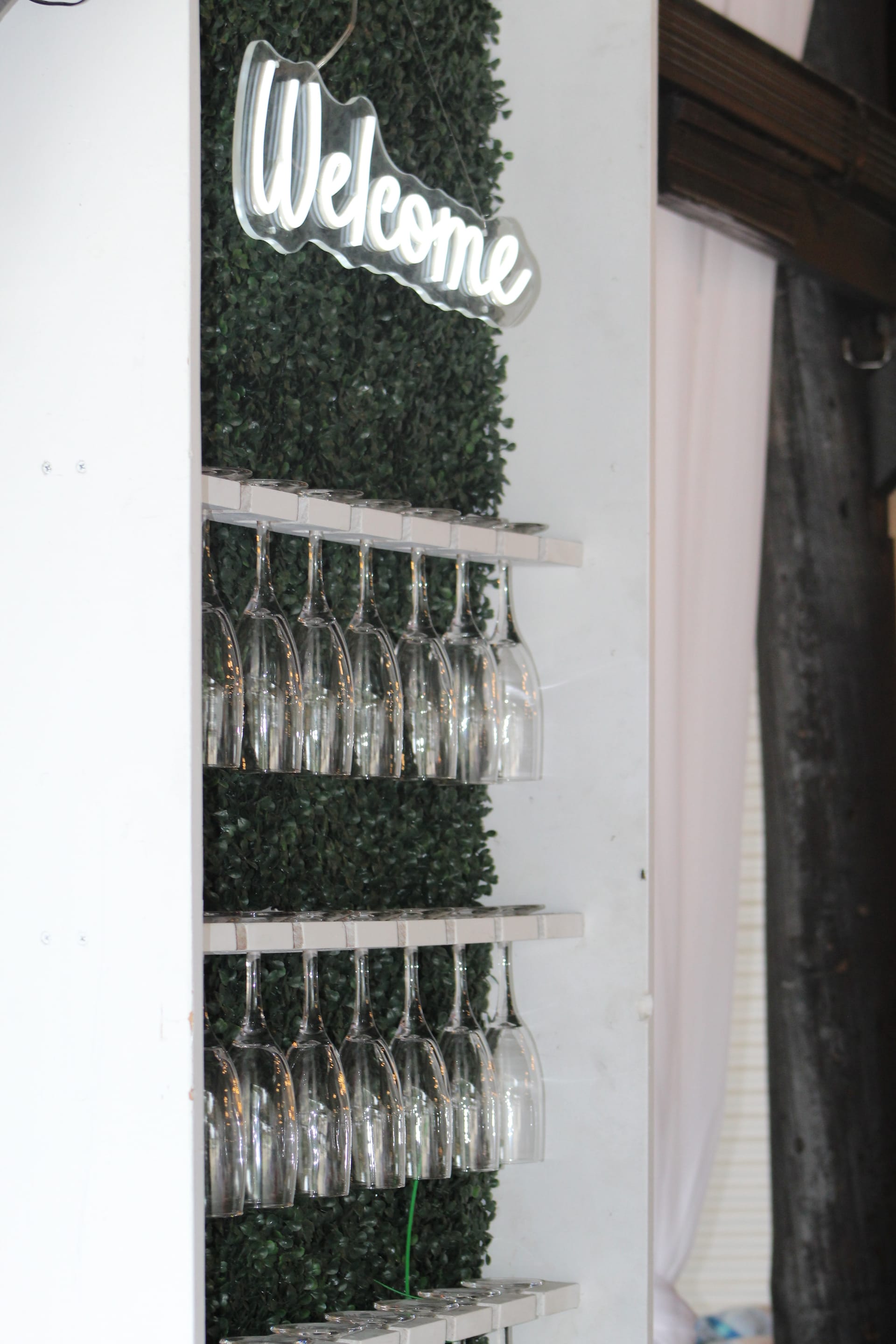Rows of stemmed wine glasses hanging upside down on a white rack mounted against a green boxwood wall beneath a glowing neon Welcome sign at Scarborough House wedding venue.