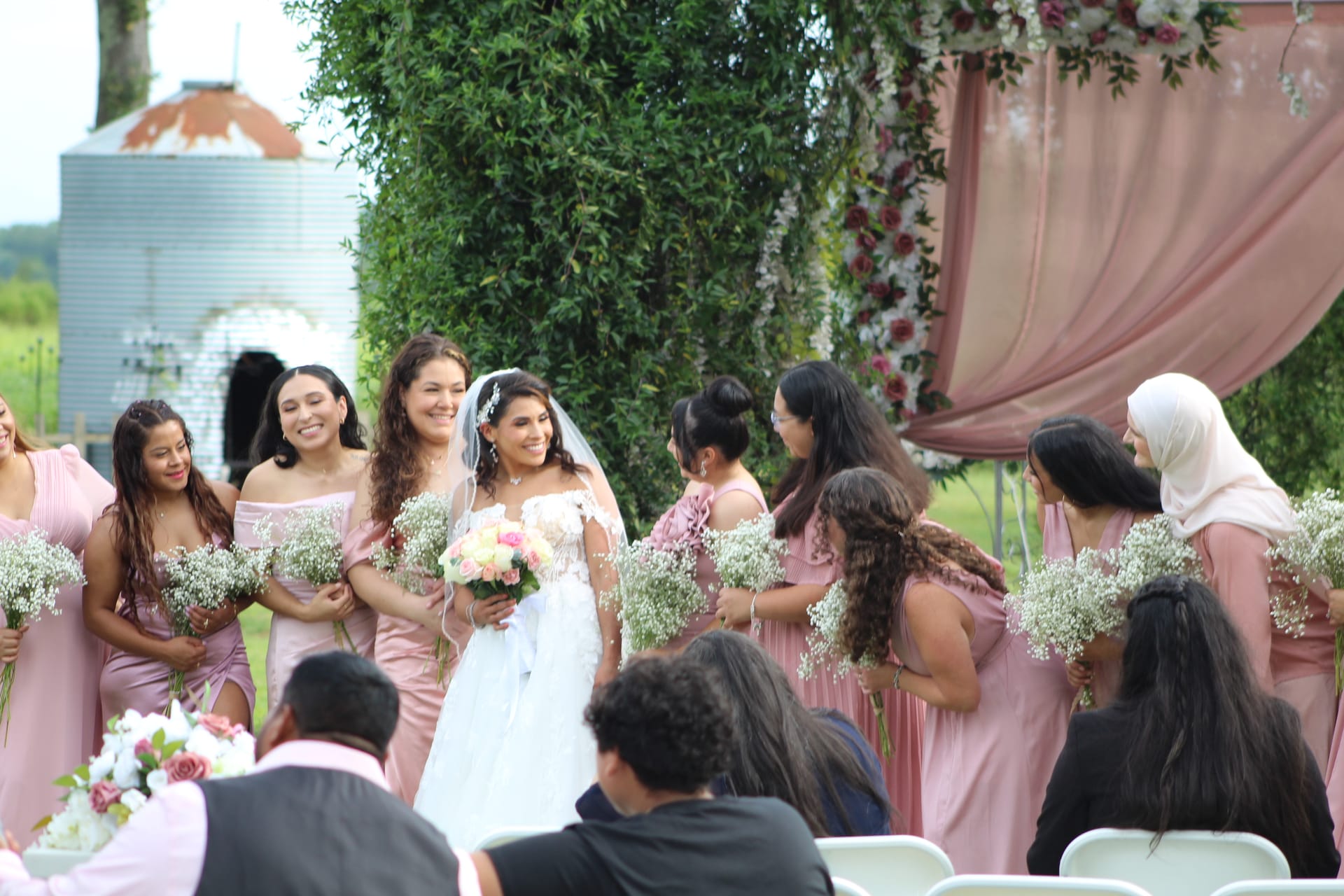 Smiling bride in a white gown and veil walks with her bridesmaids in dusty-rose dresses holding baby's breath bouquets during an outdoor ceremony at a lush garden venue.