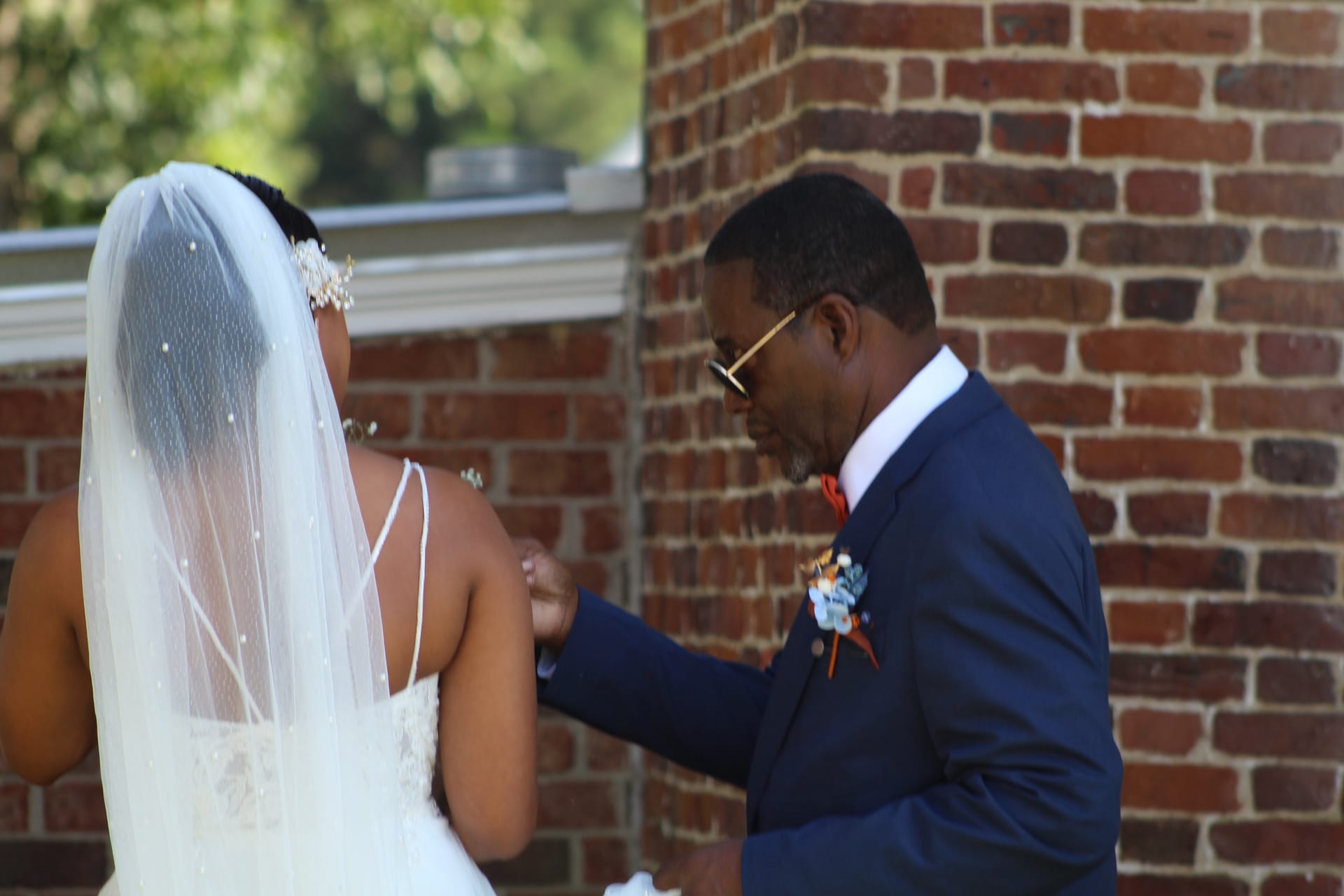 A man in a navy suit with a boutonniere tenderly adjusts the back of a bride's white gown outside a historic brick building on her wedding day.