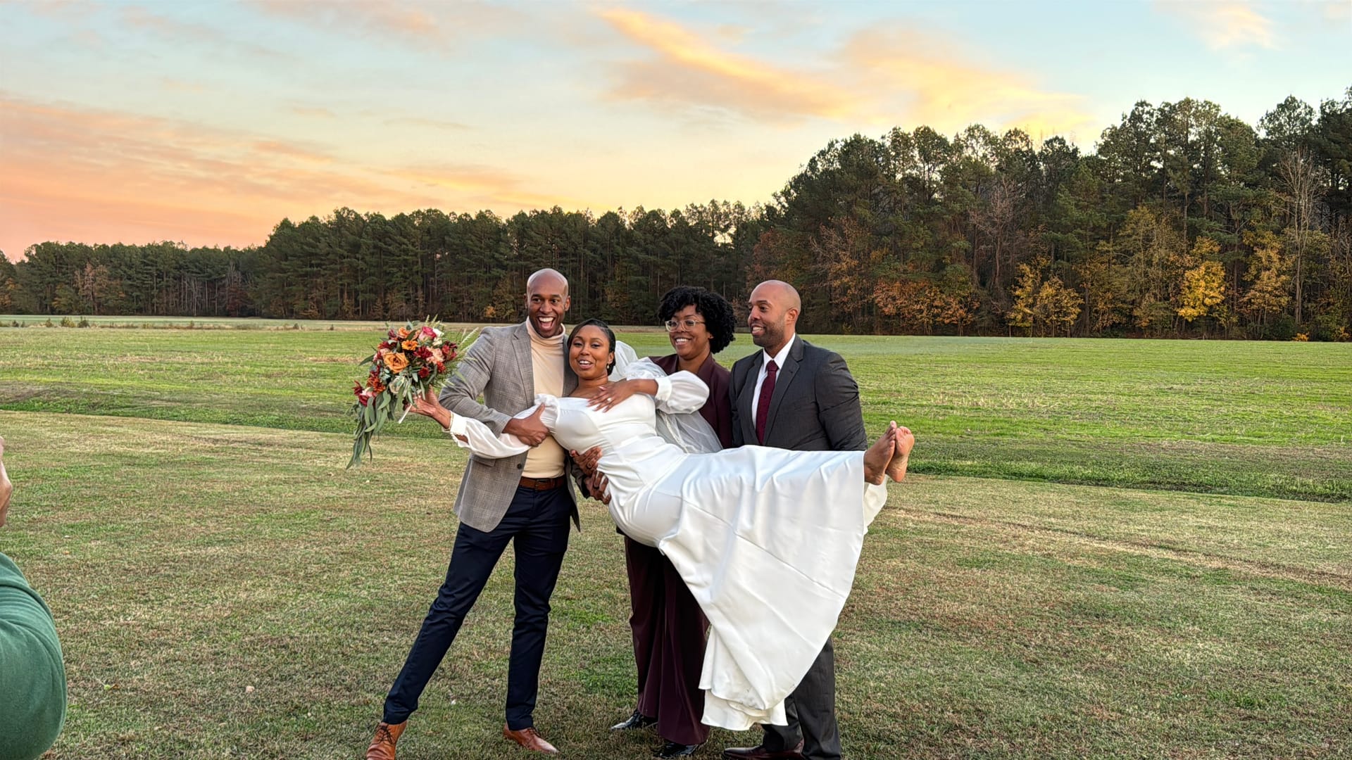 Bride in a white gown is lifted by two groomsmen while a bridesmaid stands beside them smiling, all set against a vibrant autumn sunset over open farmland fields at Scarborough House.