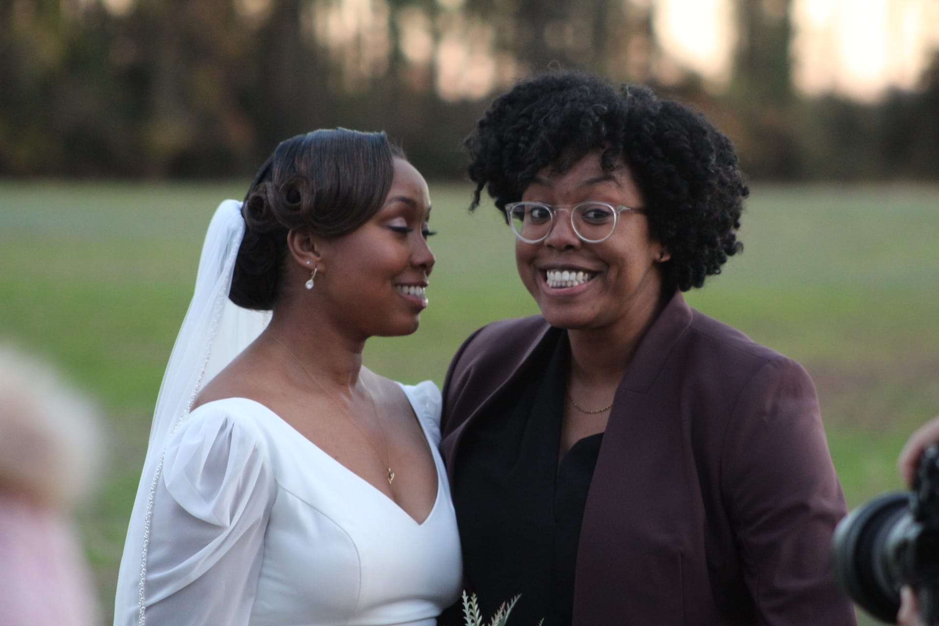 A smiling bride in a white gown and veil shares a joyful moment with her partner in a burgundy blazer against a softly blurred field and tree backdrop at Scarborough House.