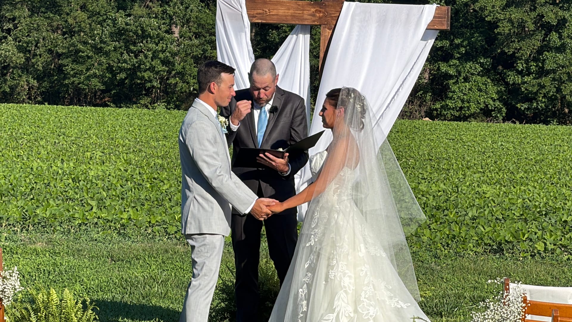 A bride in a lace gown and veil holds hands with her groom in a light gray suit as an officiant reads from a book beneath a draped wooden arch surrounded by green farmland fields.
