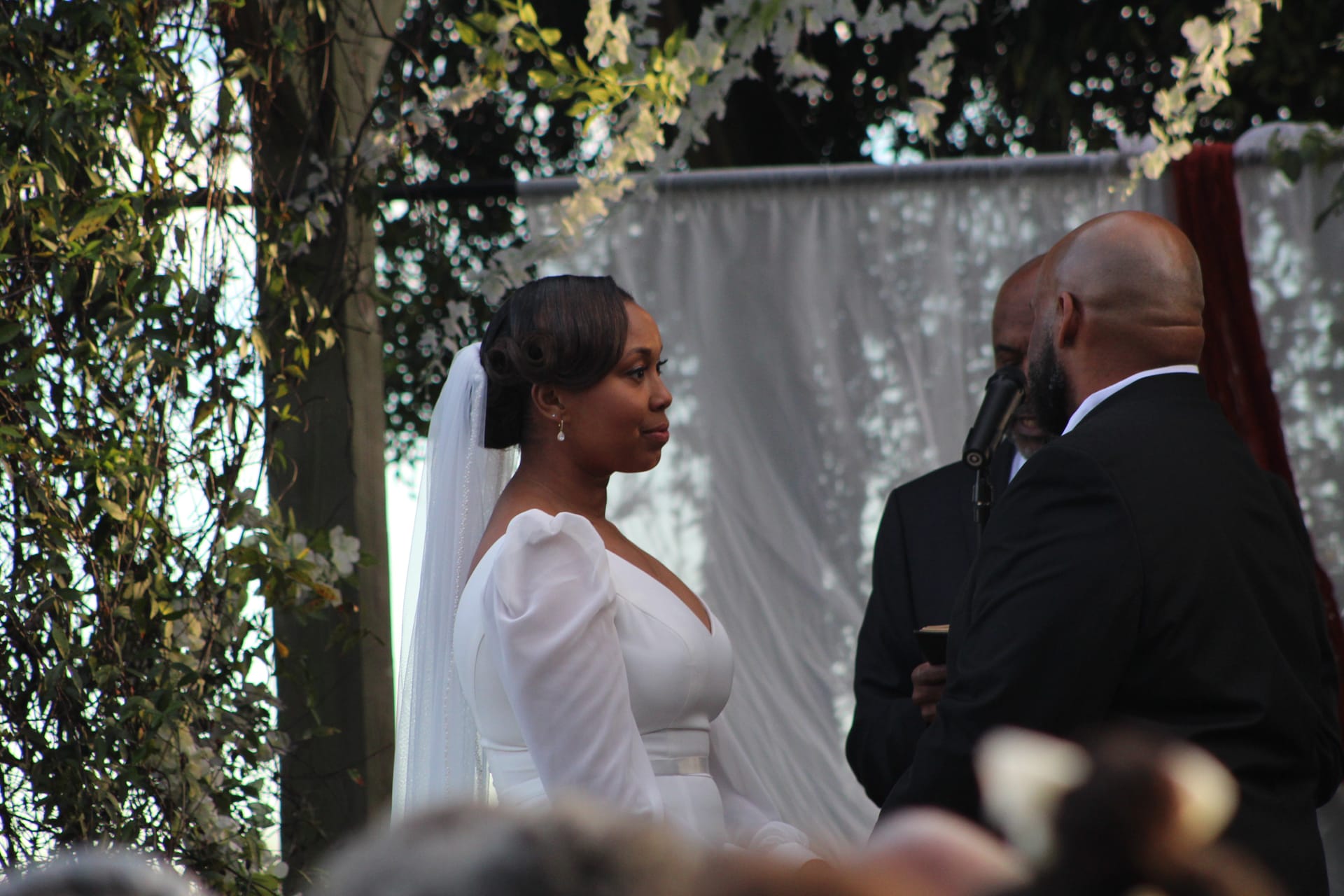 Bride in a long-sleeve white gown and sheer veil gazes at her groom during an outdoor wedding ceremony beneath a lush floral arch and draped fabric backdrop.