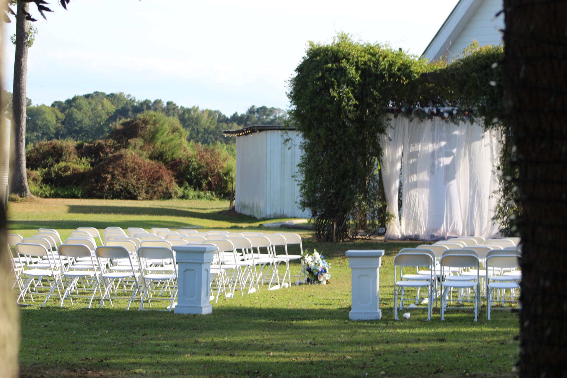 Outdoor wedding ceremony setup at Scarborough House featuring rows of white folding chairs flanking a center aisle with stone pedestals, blue floral arrangements, and a white draped arbor covered in lush greenery.
