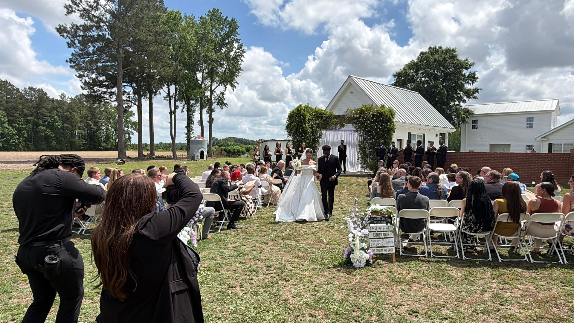 Bride walking down the outdoor aisle toward a floral arch at Scarborough House estate ceremony lawn, surrounded by seated guests with the historic white farmhouse in the background.