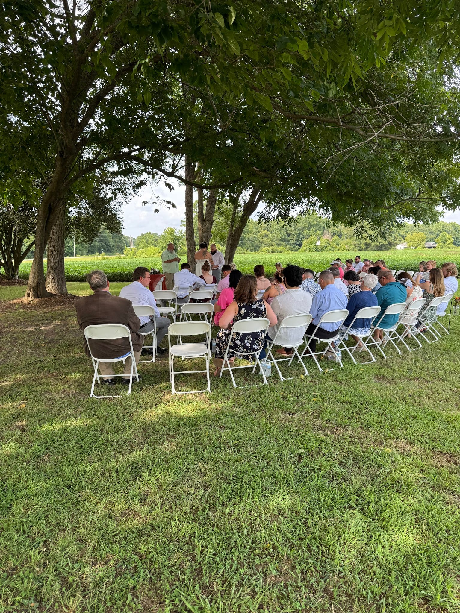Guests seated in white folding chairs on the ceremony lawn at Scarborough House watch an intimate outdoor wedding ceremony beneath mature oak trees with green farmland fields in the background.