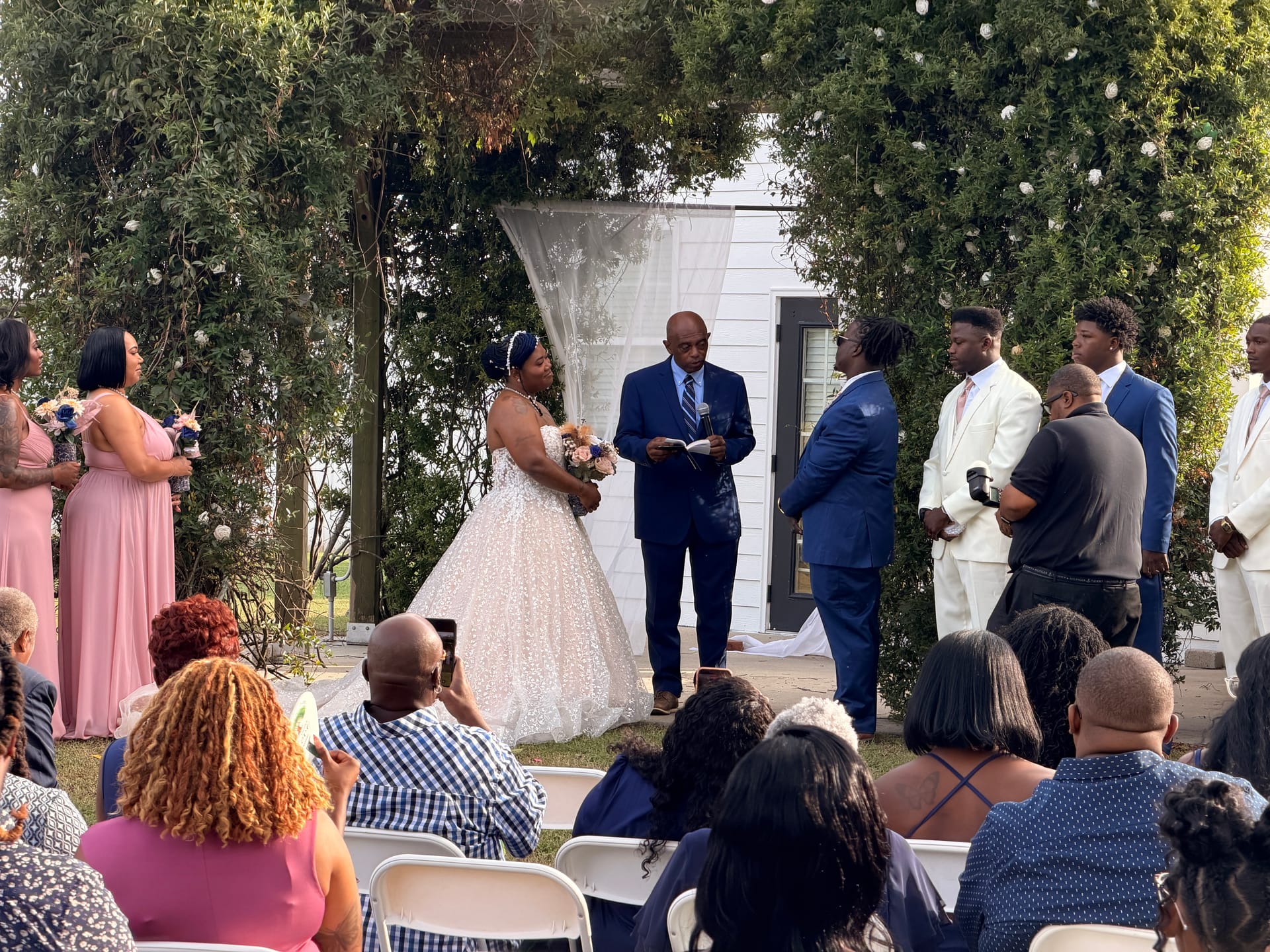 Bride in a sparkling ball gown stands at the altar beneath a lush floral arch during an outdoor wedding ceremony at Scarborough House, surrounded by bridesmaids in pink and groomsmen in cream and navy suits.