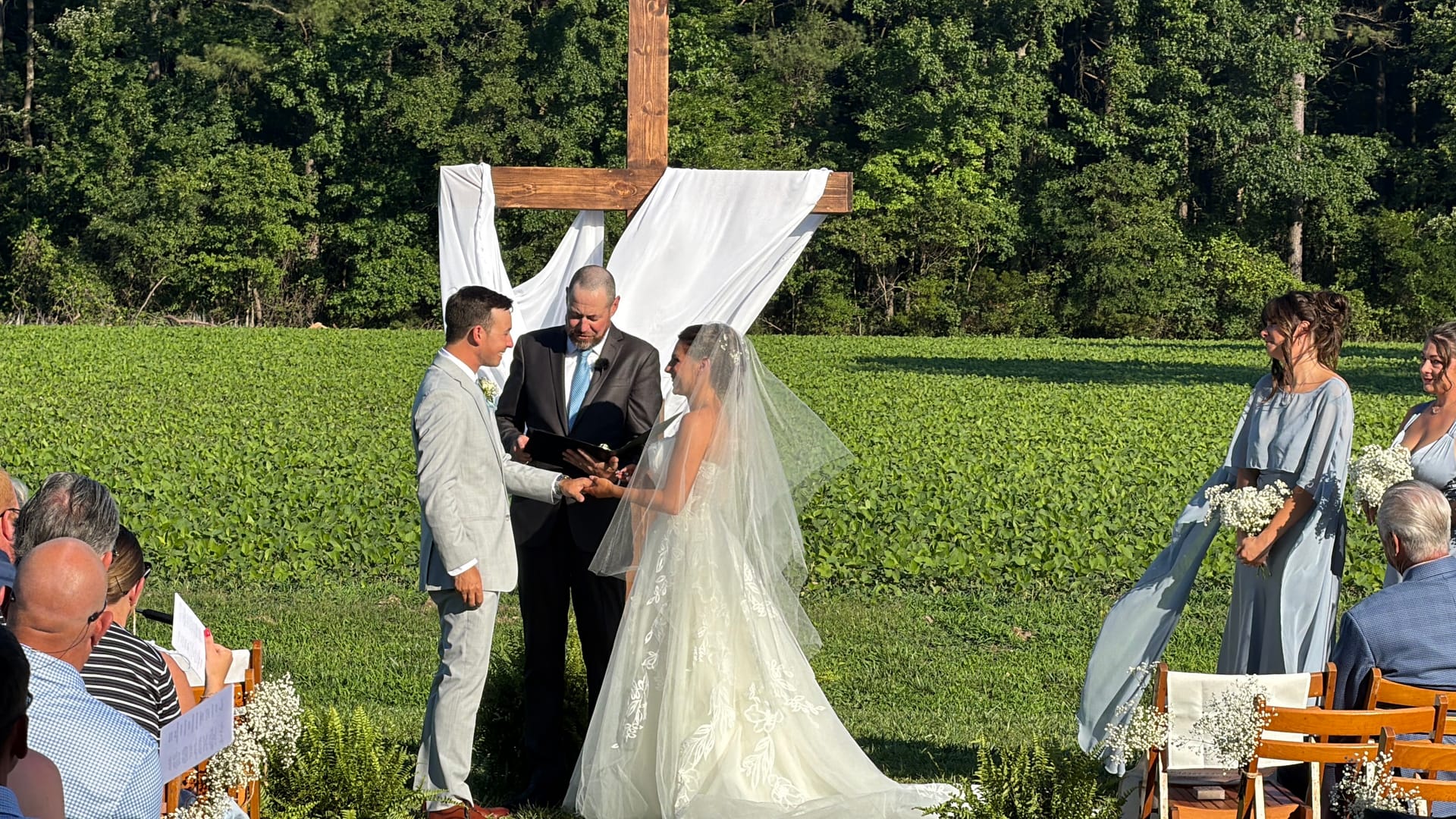 Bride and groom exchange vows at an outdoor ceremony on the estate lawn beneath a rustic wooden cross draped in white fabric, surrounded by lush green fields and wooded scenery.