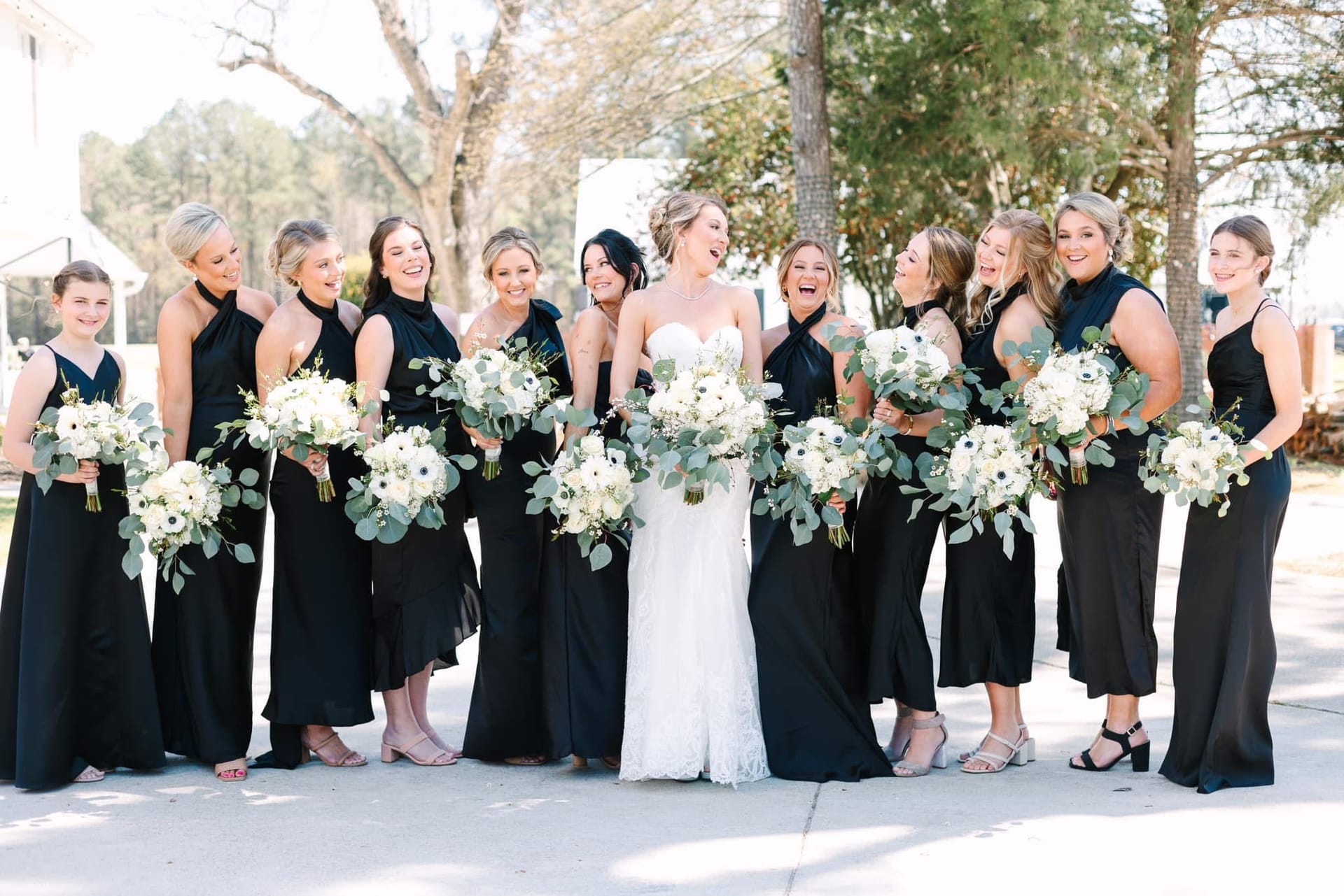 Bride in a white lace gown laughing with eleven bridesmaids in black dresses holding white floral bouquets with eucalyptus greenery outdoors at Scarborough House.