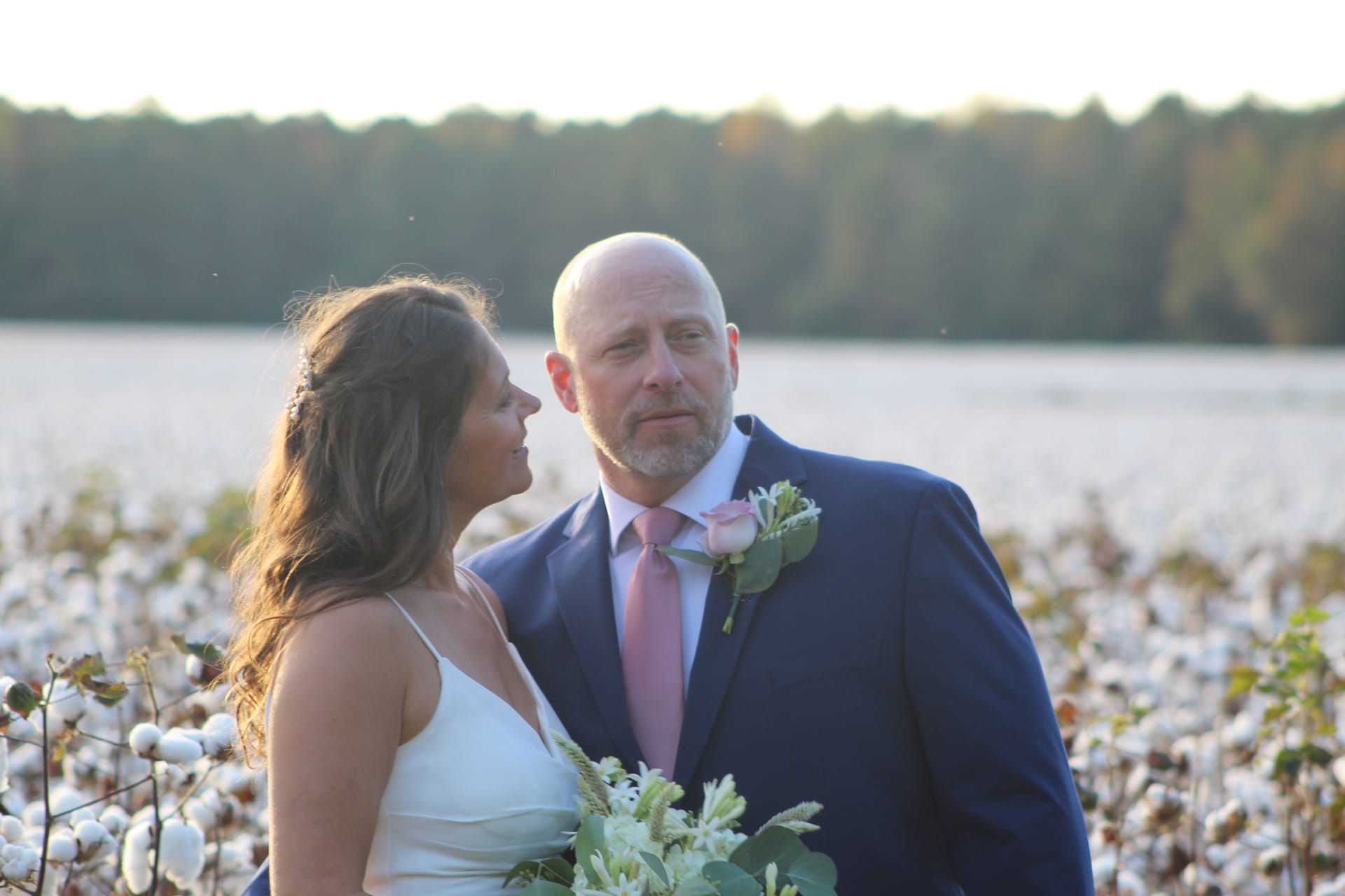 Bride in white gown gazing lovingly at groom in navy suit with pink boutonniere as they stand together in a sunlit cotton field at golden hour.