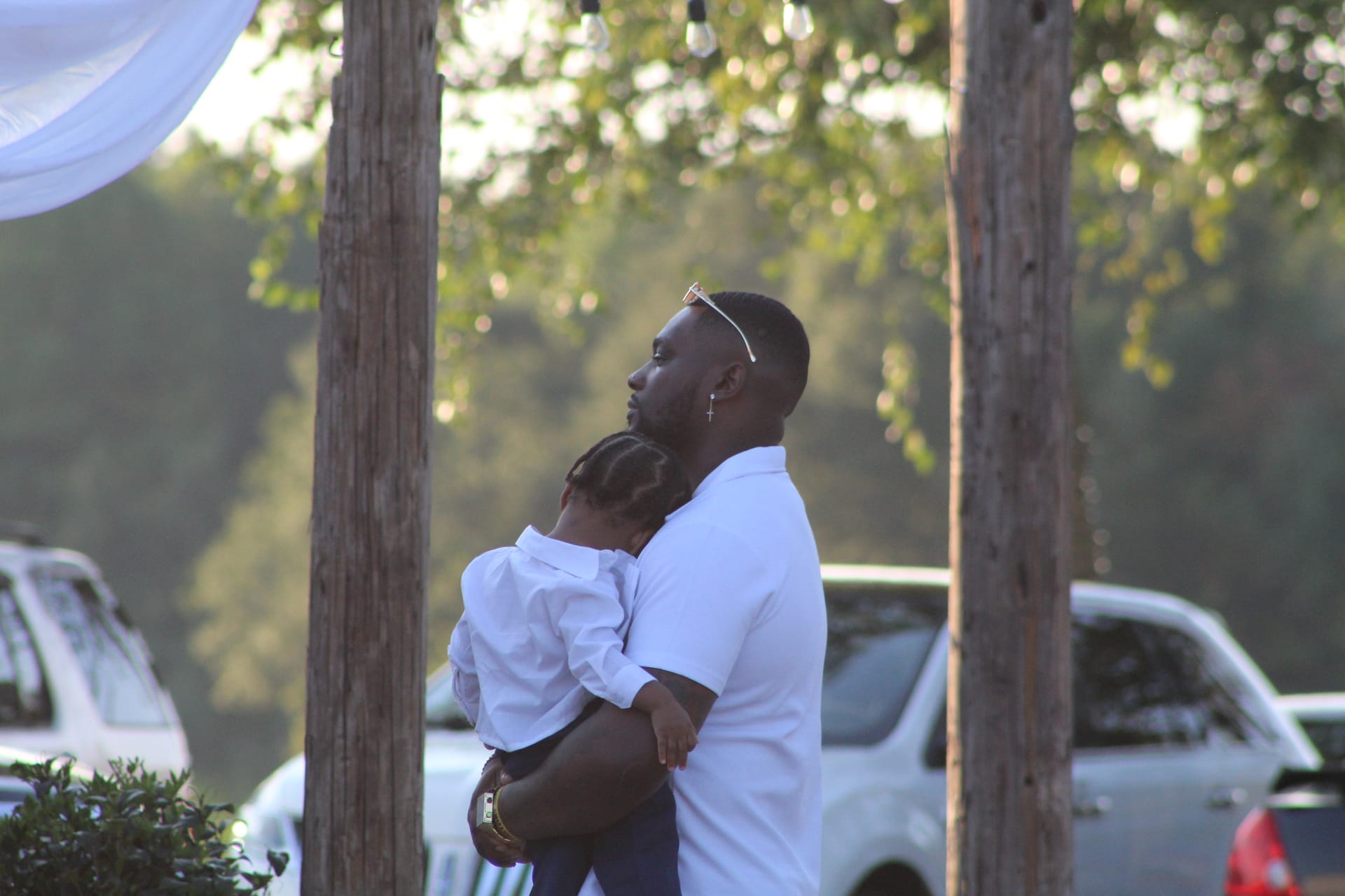A man in a white polo shirt holding a young child dressed in white during an outdoor wedding celebration beneath rustic wooden posts and string lights at Scarborough House estate.