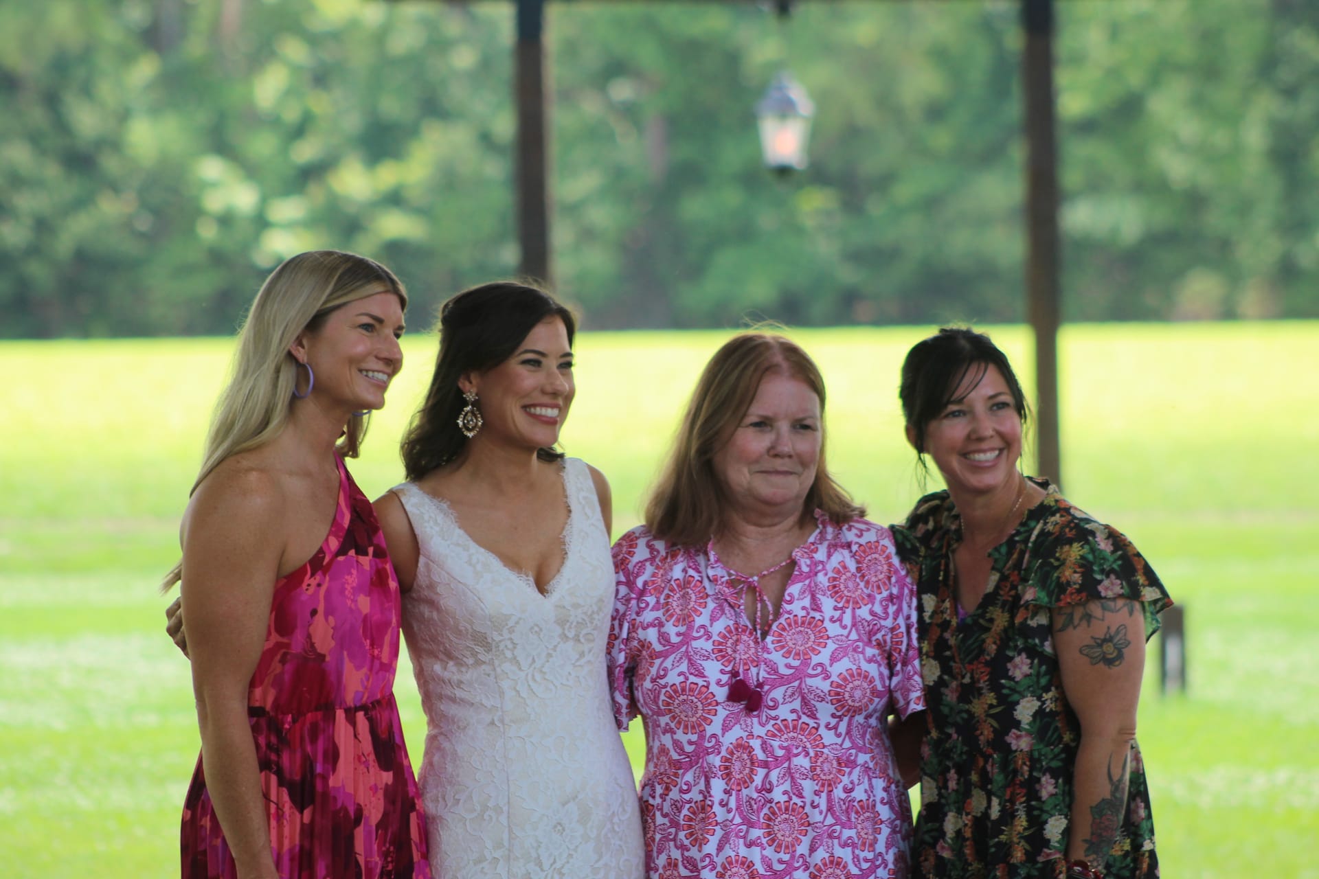 Bride in a white lace gown smiles alongside three guests in floral and pink dresses on the verdant ceremony lawn at Scarborough House.