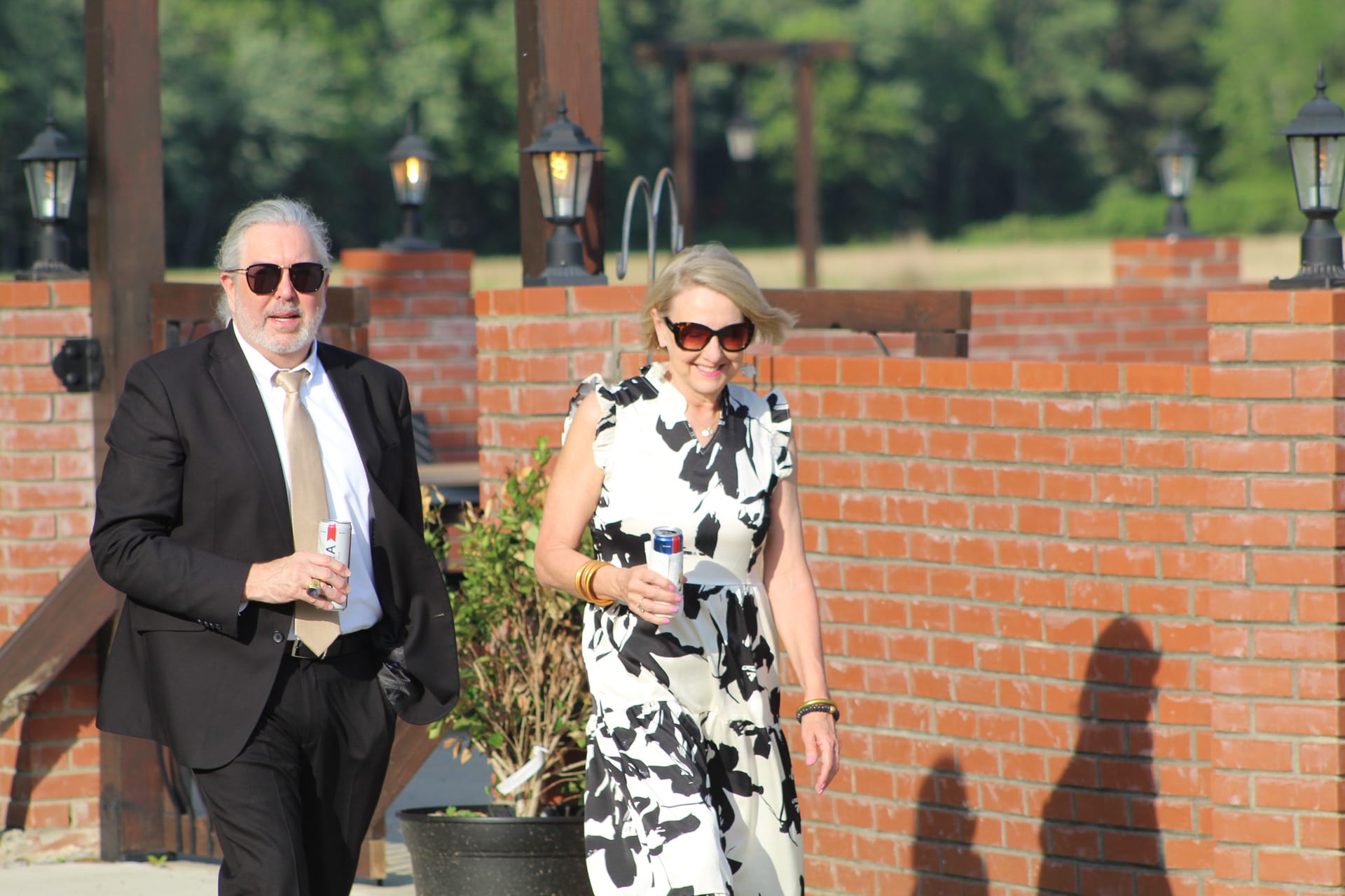 A smiling couple dressed in formal attire walks along a brick wall pathway lined with lantern posts at Scarborough House estate wedding venue.