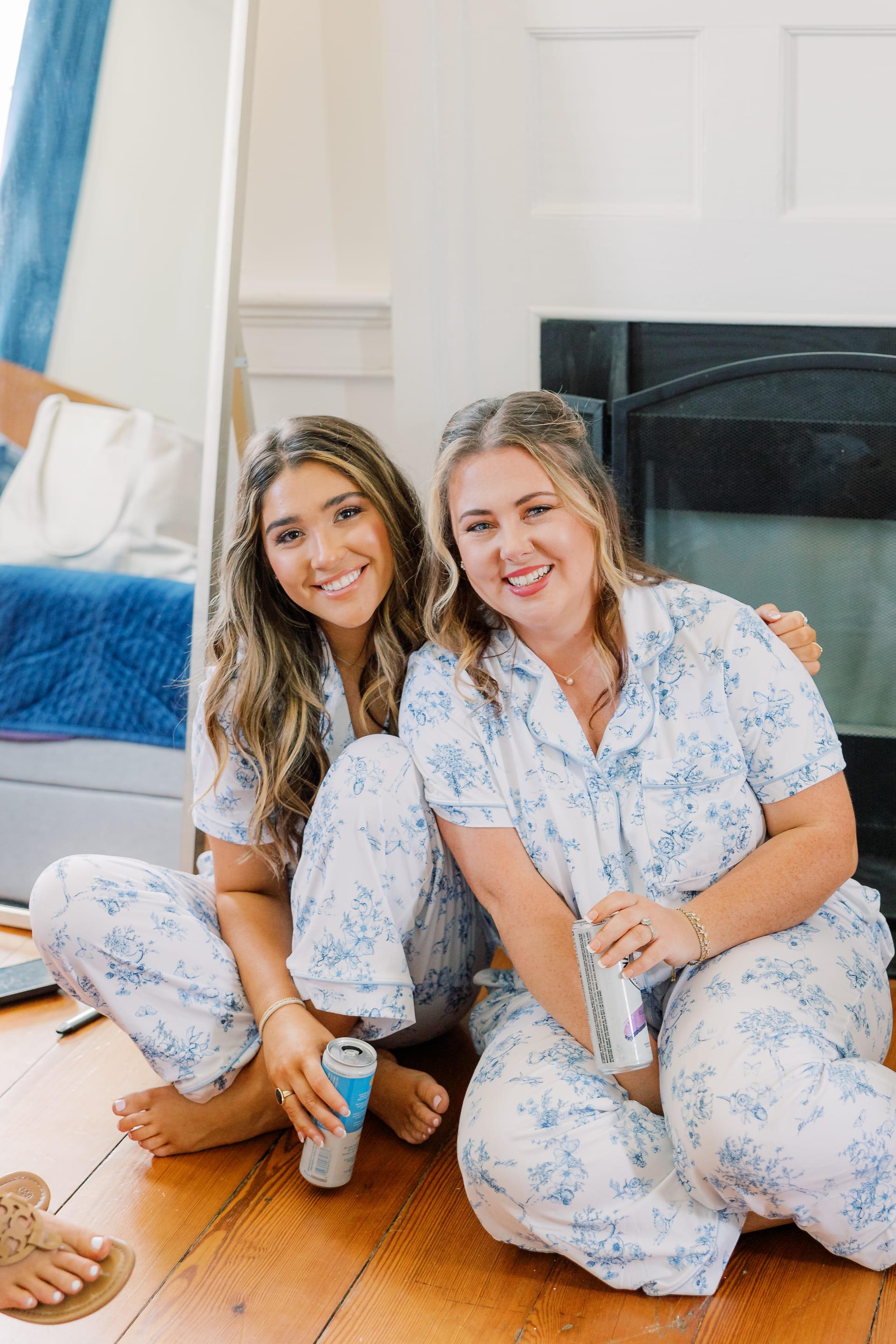 Two bridesmaids in matching blue floral pajamas sitting on a hardwood floor smiling and holding canned drinks during wedding morning preparations.