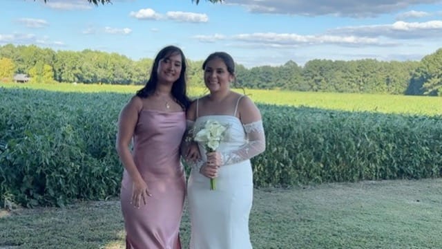 A bride in a white off-shoulder gown holding a white floral bouquet poses with a guest in a blush satin dress in front of green farmland fields and blue skies at Scarborough House.