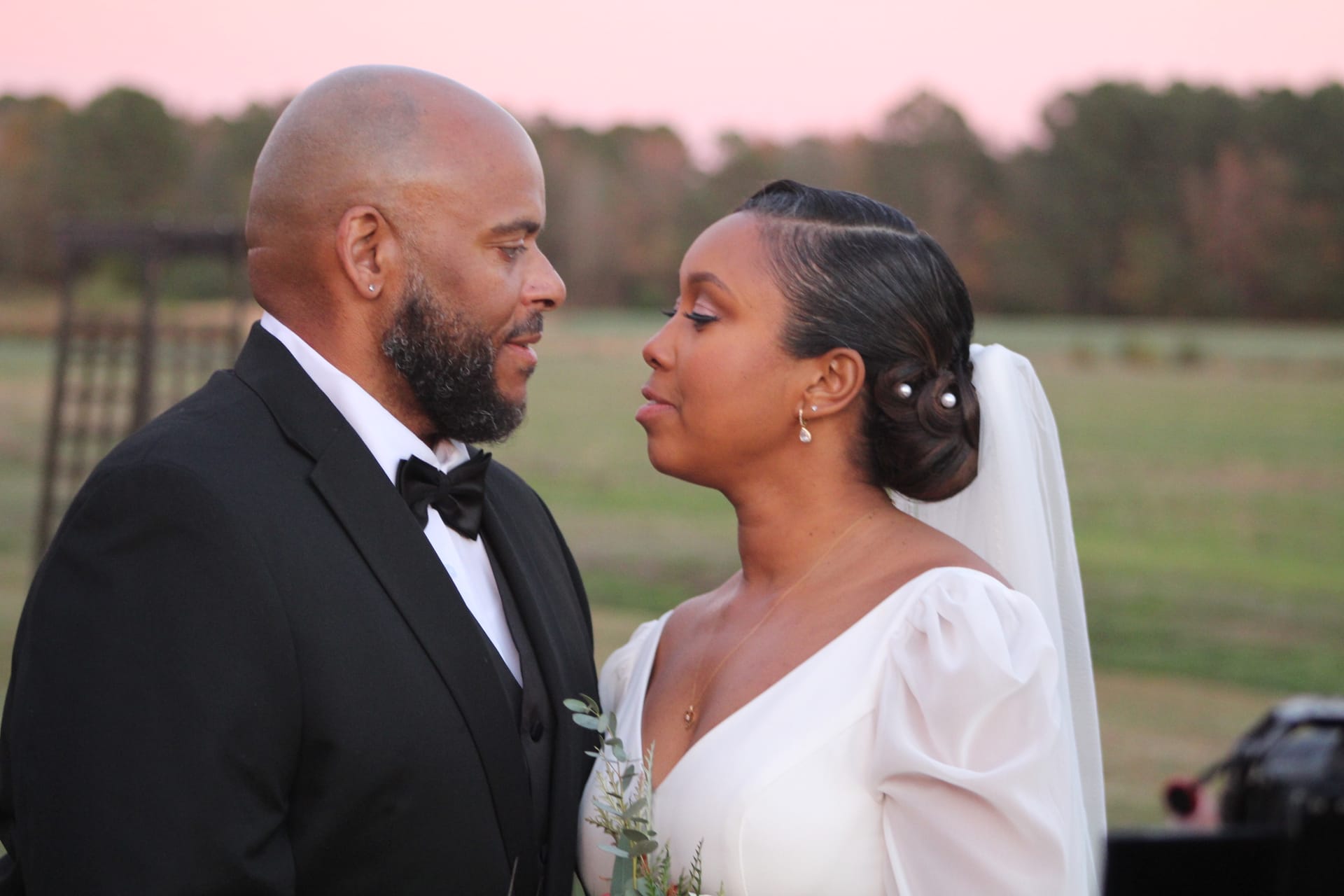 Bride in a white gown with pearl hair pins and groom in a black tuxedo sharing an intimate gaze at golden hour with open farmland fields and a soft pink sky behind them at Scarborough House.