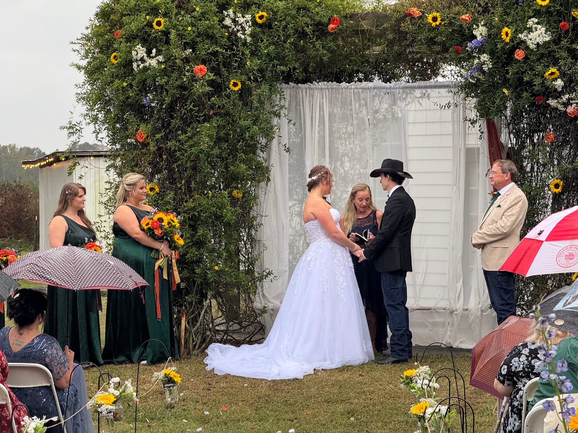 Bride in a white ballgown exchanges vows with her groom in a black cowboy hat beneath a lush floral arch decorated with sunflowers and greenery on the ceremony lawn at Scarborough House, surrounded by bridesmaids in emerald green and wedding guests holding umbrellas on a cloudy day.